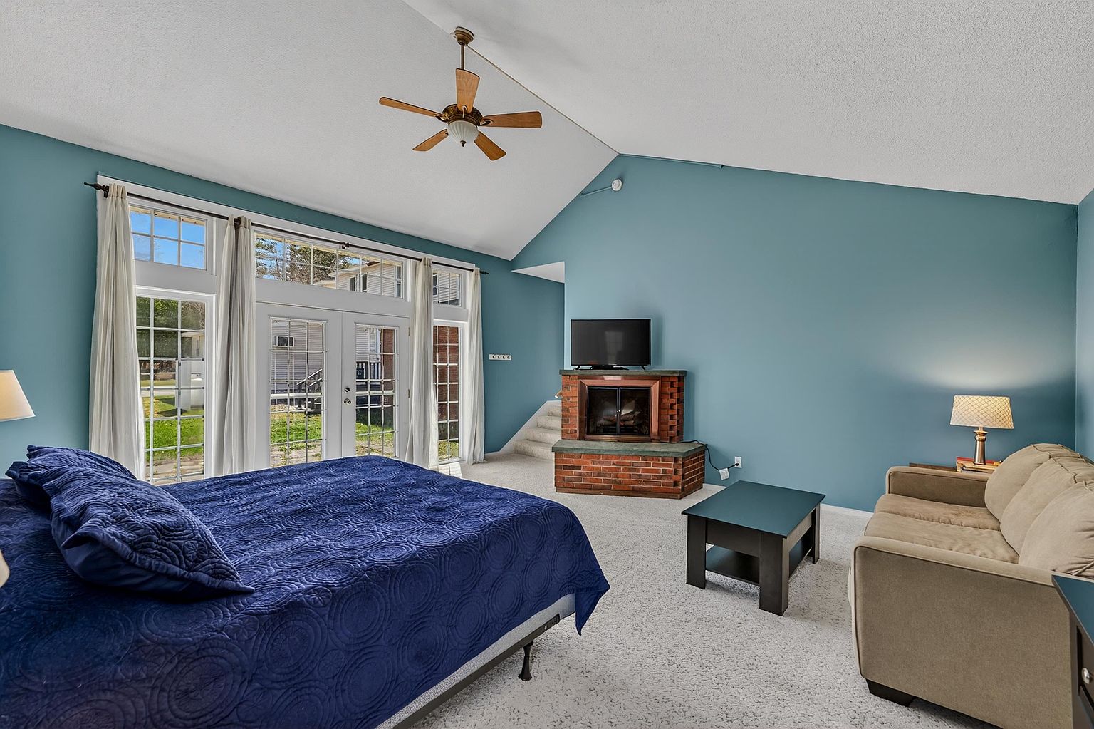 This spacious primary bedroom features a vaulted ceiling and a large set of glass doors that provide abundant natural light and a view of the outdoors. The room is anchored by a brick fireplace with a hearth, positioned near a set of stairs, and includes a comfortable seating area with a beige sofa. The blue walls and neutral carpeting create a calm, versatile atmosphere suitable for a primary suite.