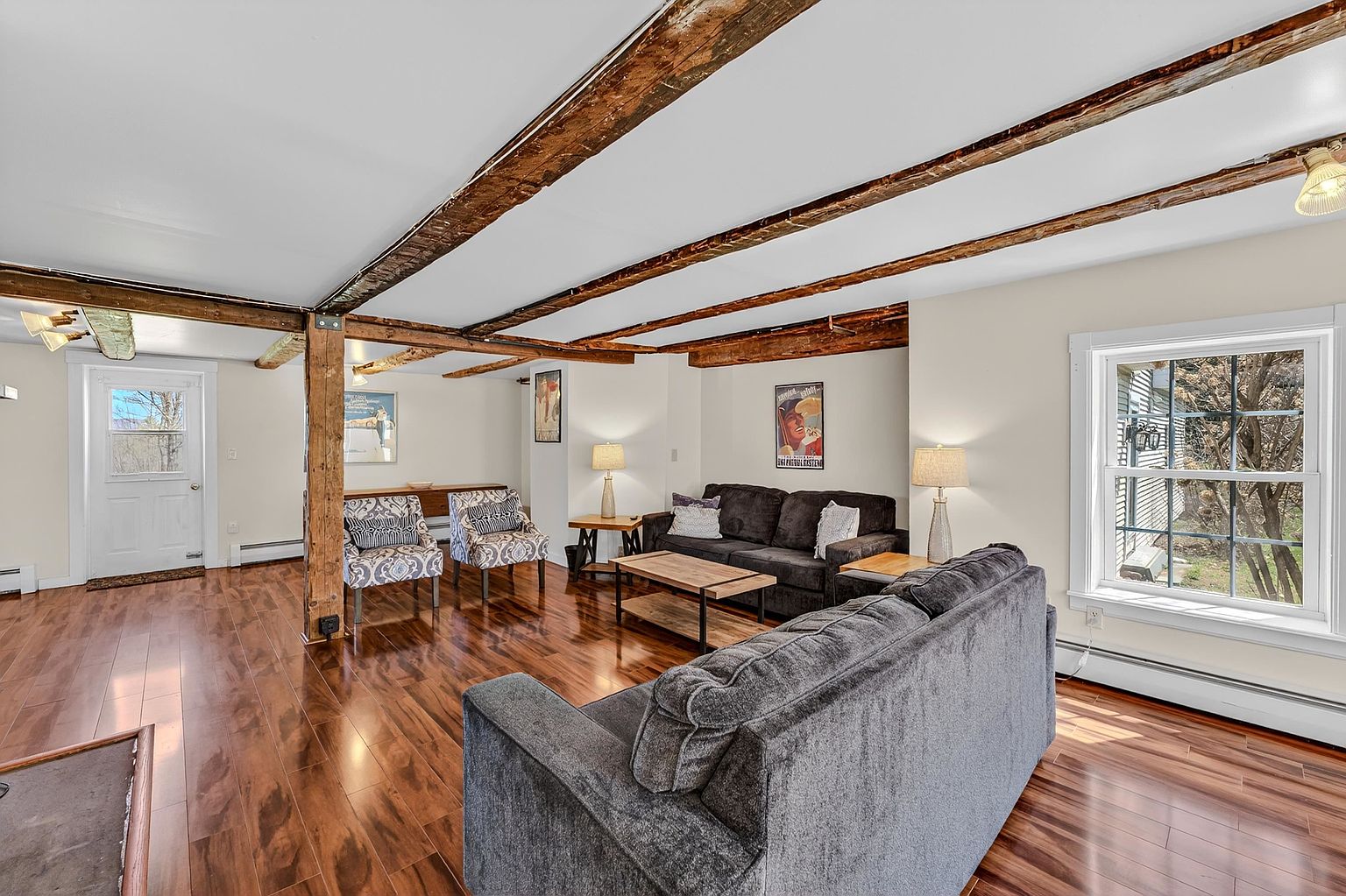 This spacious living room features warm-toned wood flooring and exposed wooden ceiling beams, creating a rustic and inviting atmosphere. The room is furnished with two dark gray sofas and a pair of patterned accent chairs, centered around a wooden coffee table. Large windows and a glass-paneled door allow for ample natural light, while framed artwork and table lamps add a personalized touch to the space.