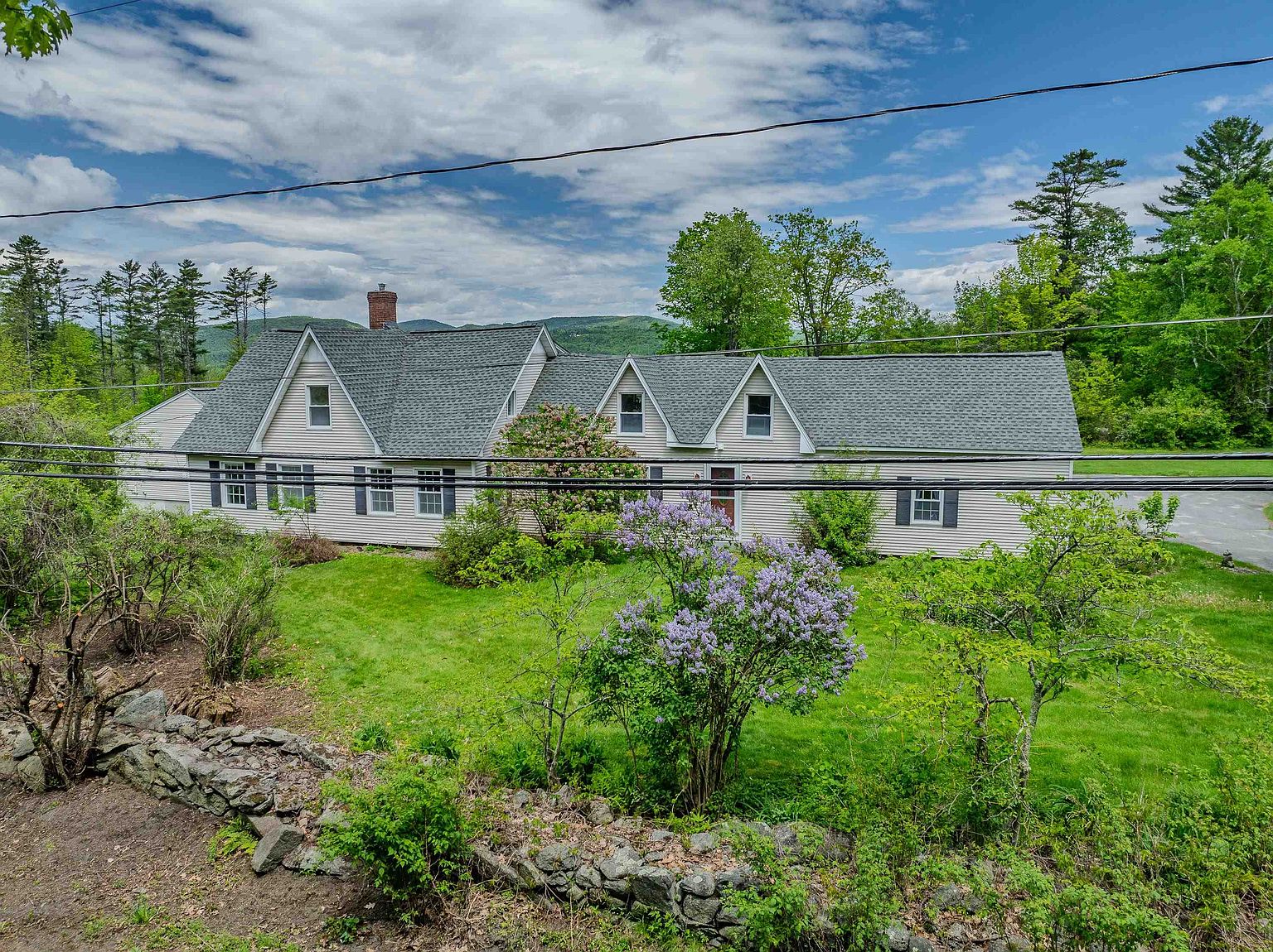 This image captures a wide, eye-level view of a traditional, single-story suburban home with light-colored horizontal siding and a dark shingled roof. The house features multiple gabled dormers and a central entryway, set against a backdrop of lush green trees and rolling hills under a bright, cloudy sky. A well-maintained lawn with a blooming lilac bush in the foreground and a rustic stone wall adds to the property's charming, rural aesthetic.