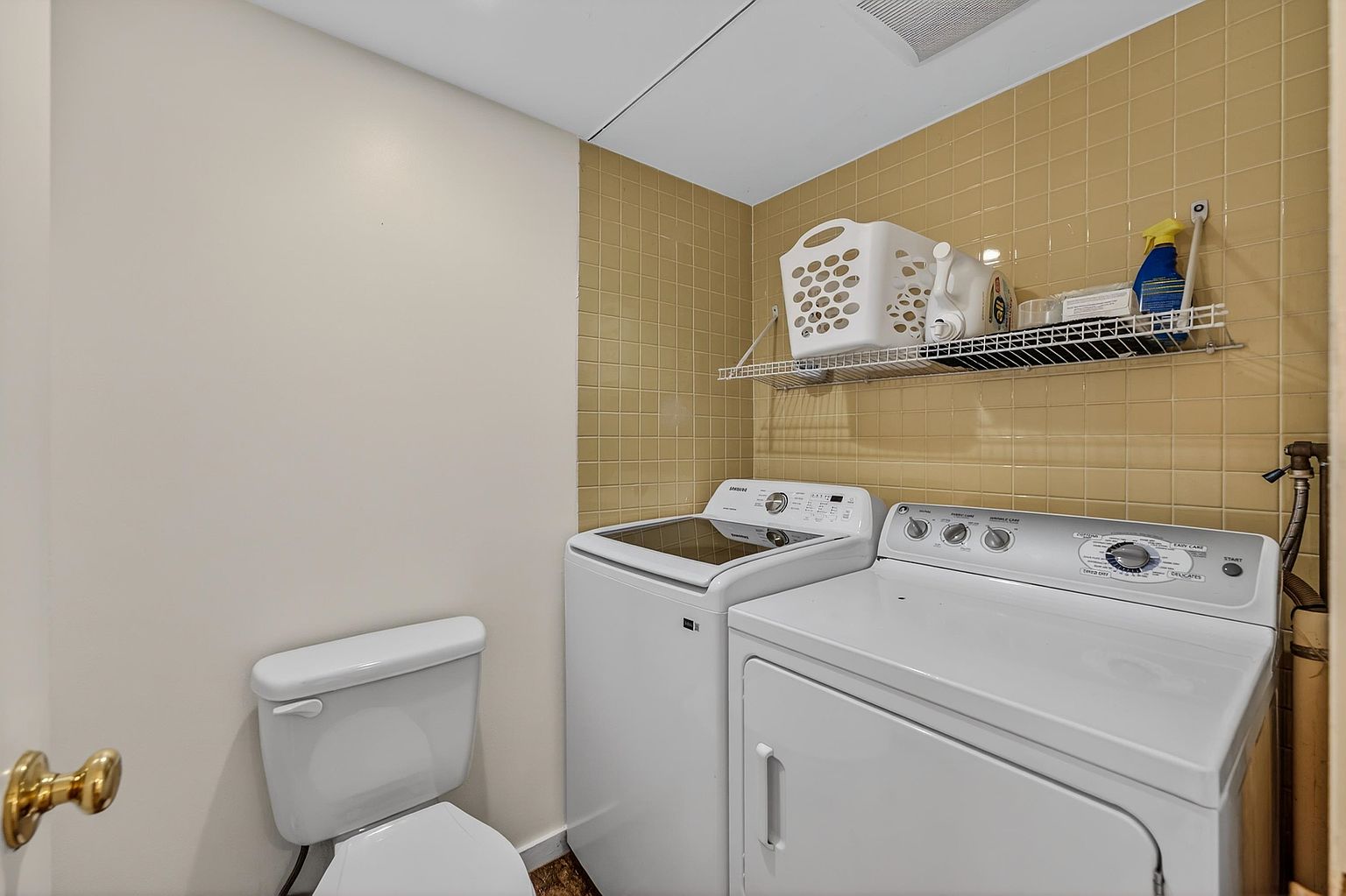 This functional laundry room features a side-by-side washer and dryer set positioned against a wall tiled with light-colored square tiles. A wire shelving unit mounted above the appliances provides convenient storage for laundry supplies, while a white toilet is situated to the left, indicating a combined utility and bathroom space. The room is brightly lit and offers a practical, compact layout.