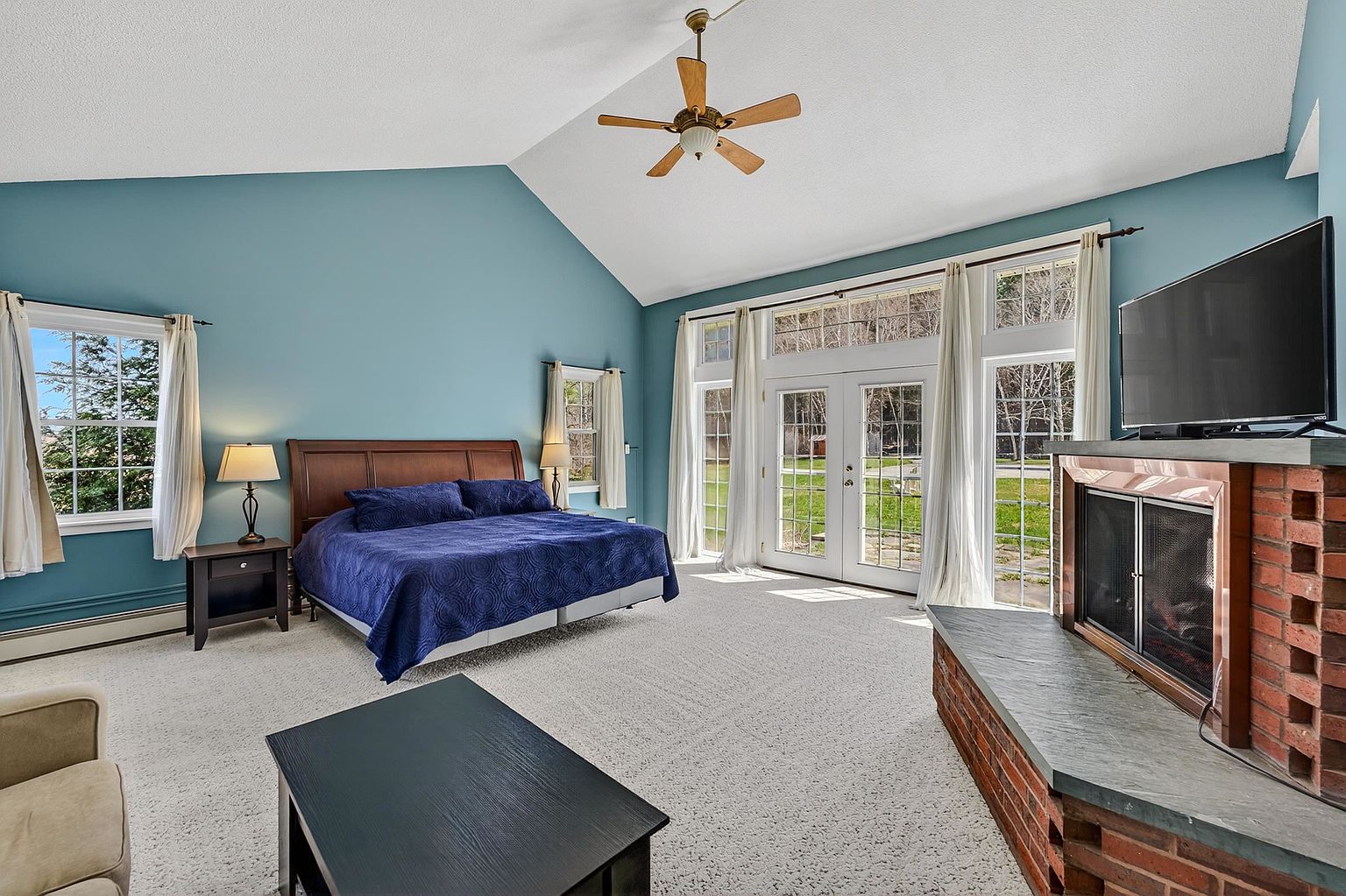This spacious primary bedroom features vaulted ceilings, light blue walls, and plush carpeting throughout. A large bed with a dark wood frame serves as the focal point, flanked by two nightstands with lamps, while a brick fireplace with a stone hearth and a mounted television adds warmth to the room. The space is brightened by large windows and glass doors that provide a view of the outdoor greenery.