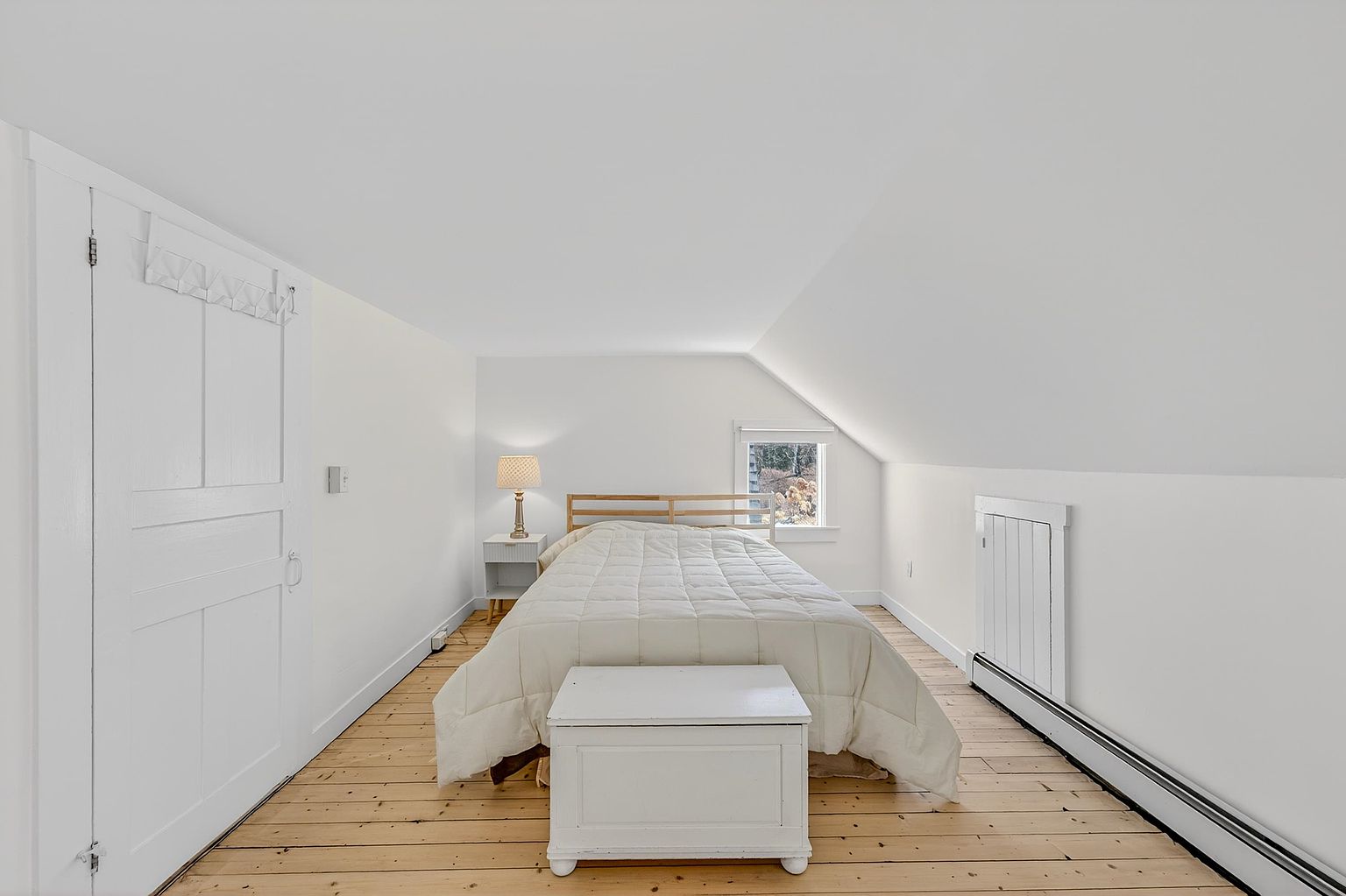 This cozy attic bedroom features a sloped ceiling and light-toned wood flooring, creating a bright and airy atmosphere. The room is furnished with a bed dressed in a white quilt, a small bedside table with a lamp, and a white storage chest at the foot of the bed. A single window provides natural light, while a white door on the left and a radiator cover on the right complete the simple, clean aesthetic.