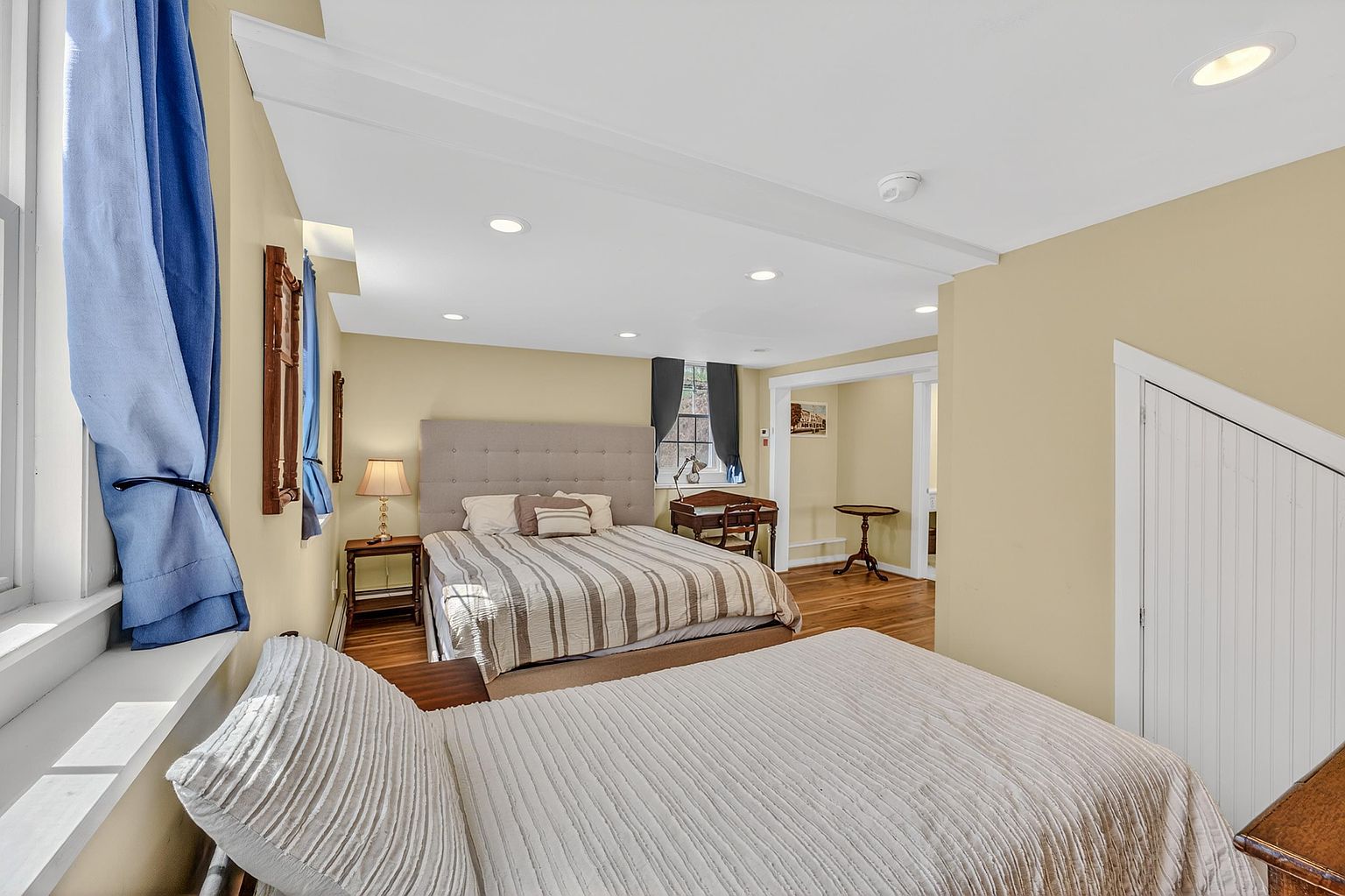 This bedroom features a dual-bed layout with a primary bed against the back wall and a second bed in the foreground. The room is painted in a soft yellow tone and includes a small wooden desk, a nightstand with a lamp, and a built-in storage closet with a sloped door. The space is illuminated by recessed lighting and natural light from the windows, creating a cozy and functional living area.