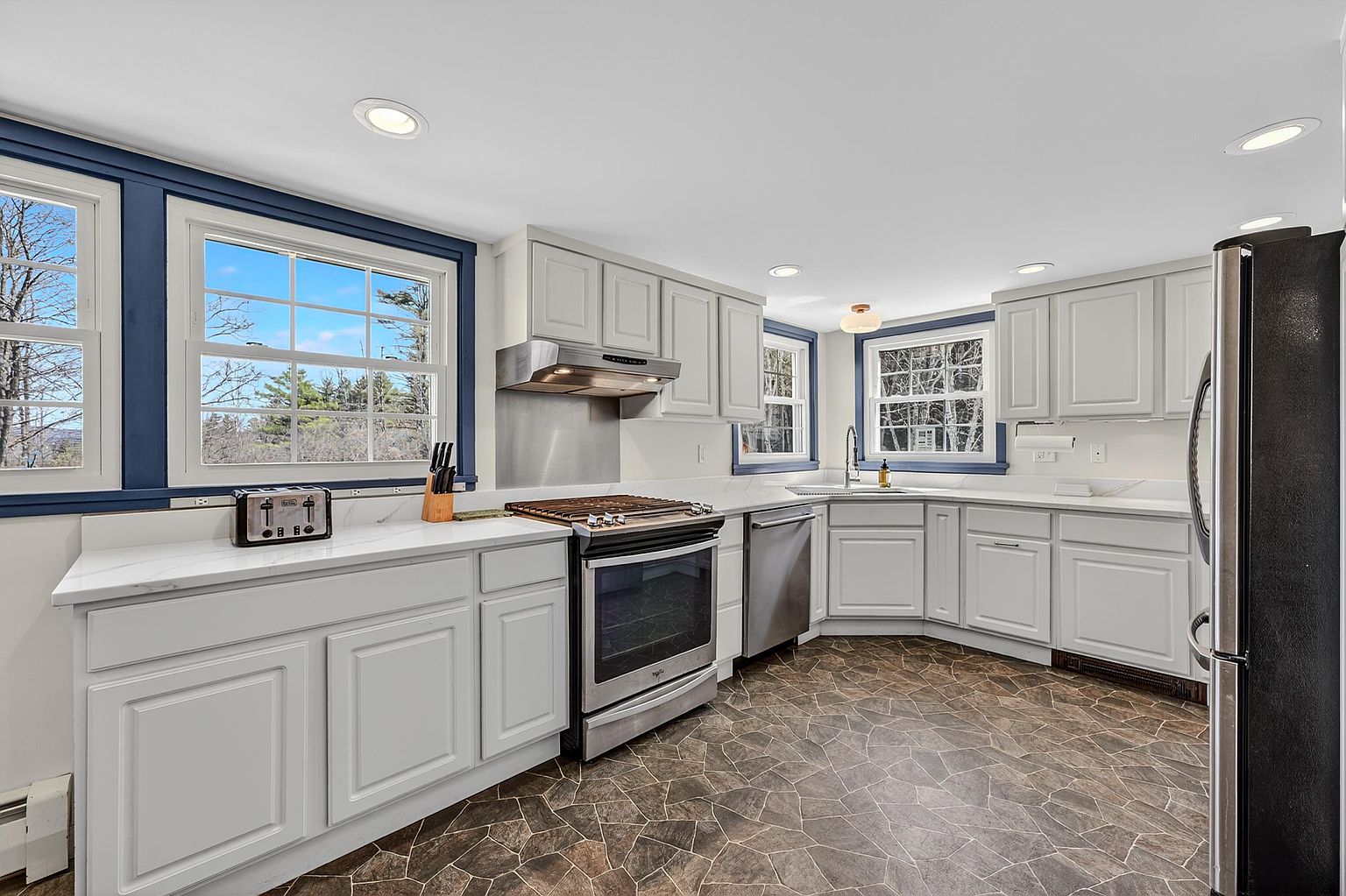 This bright and functional kitchen features light-colored cabinetry paired with white countertops and a stainless steel appliance suite. The space is anchored by a unique, multi-toned stone-look floor and is illuminated by recessed lighting and natural light streaming through multiple windows with dark blue trim. The layout provides an efficient workspace with a central range and a corner sink area, offering a clean and well-maintained aesthetic.