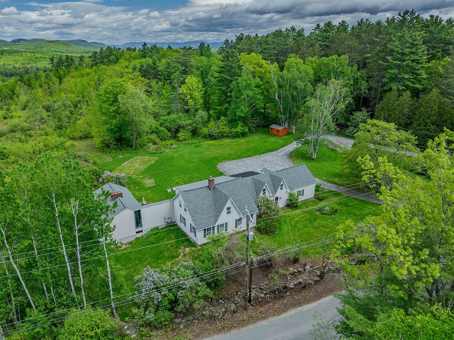 This high-angle aerial view captures a sprawling, multi-sectioned home nestled within a lush, green landscape. The property features a light-colored exterior, a gray shingled roof, and a paved driveway leading to a small shed, all surrounded by dense forest and rolling hills in the distance. The perspective highlights the expansive yard and the home's integration into its quiet, rural setting.