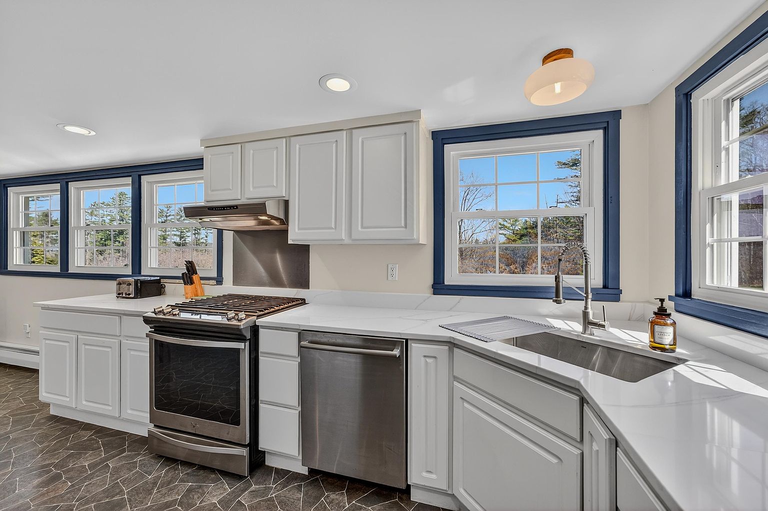 This bright, well-lit kitchen features white cabinetry paired with contrasting dark blue window trim and a unique geometric patterned floor. The space is equipped with stainless steel appliances, including a gas range and a dishwasher, set against light-colored stone countertops. Large windows provide ample natural light and views of the surrounding trees, creating an airy and inviting atmosphere.