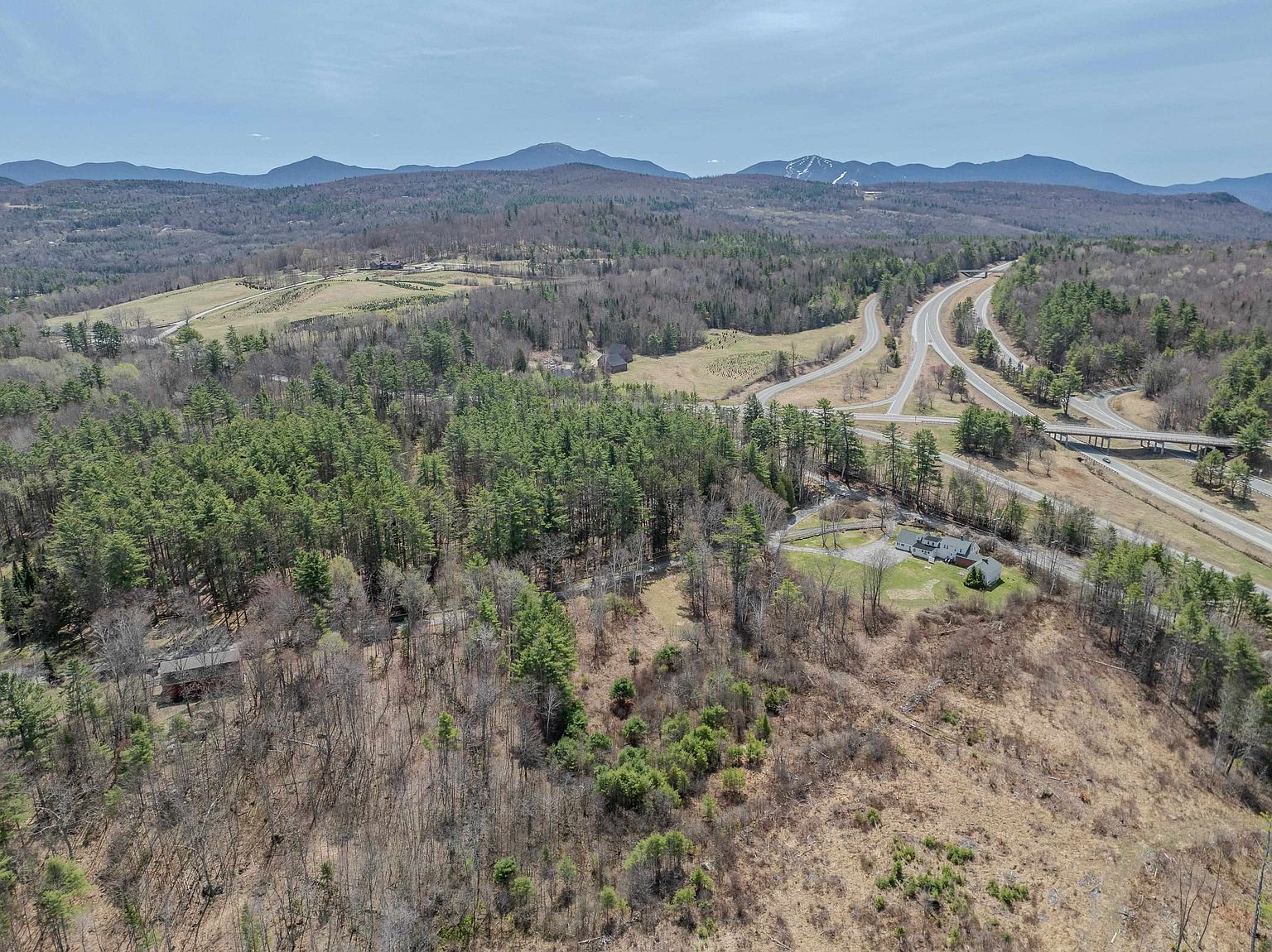 This high-angle aerial perspective captures a sprawling rural landscape featuring a residential property nestled among dense evergreen forests and open fields. A major highway interchange winds through the background, set against a backdrop of rolling mountains under a clear sky. The scene emphasizes the property's secluded location and expansive natural surroundings, highlighting its integration into the mountainous terrain.