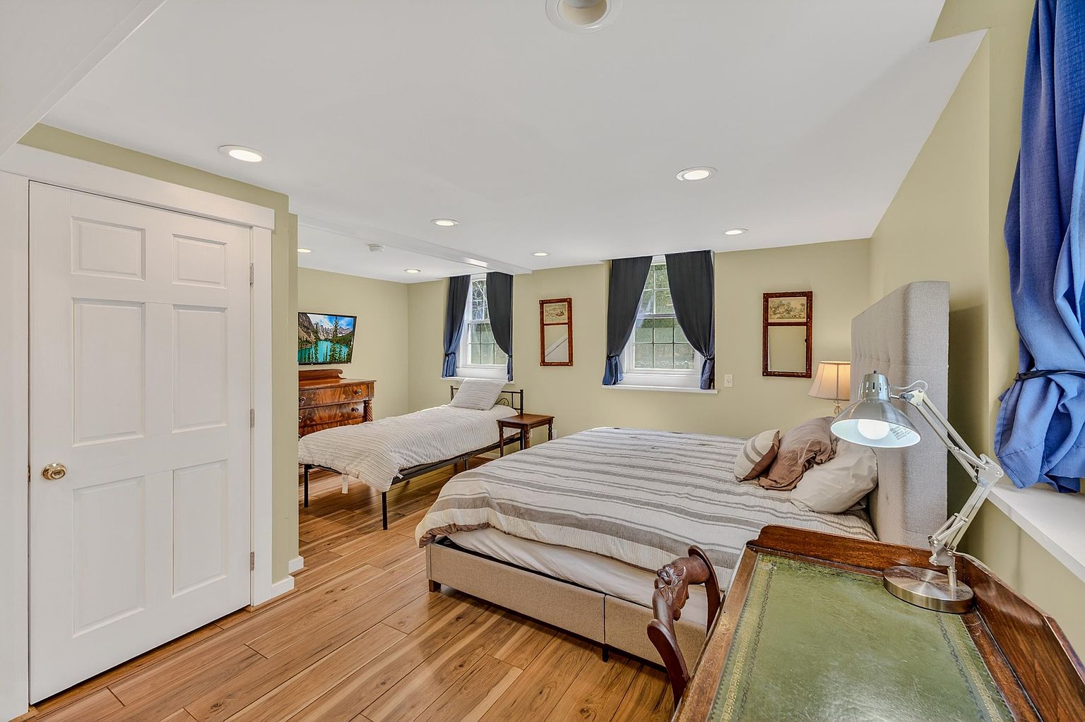 This bedroom features a multi-bed layout with a large bed in the foreground and a twin bed positioned further back, both dressed in neutral striped bedding. The room is finished with light-toned wood flooring, soft green walls, and two windows framed by blue curtains. A vintage-style desk with a green leather top and a metal task lamp sits in the immediate foreground, adding a functional workspace element to the room.