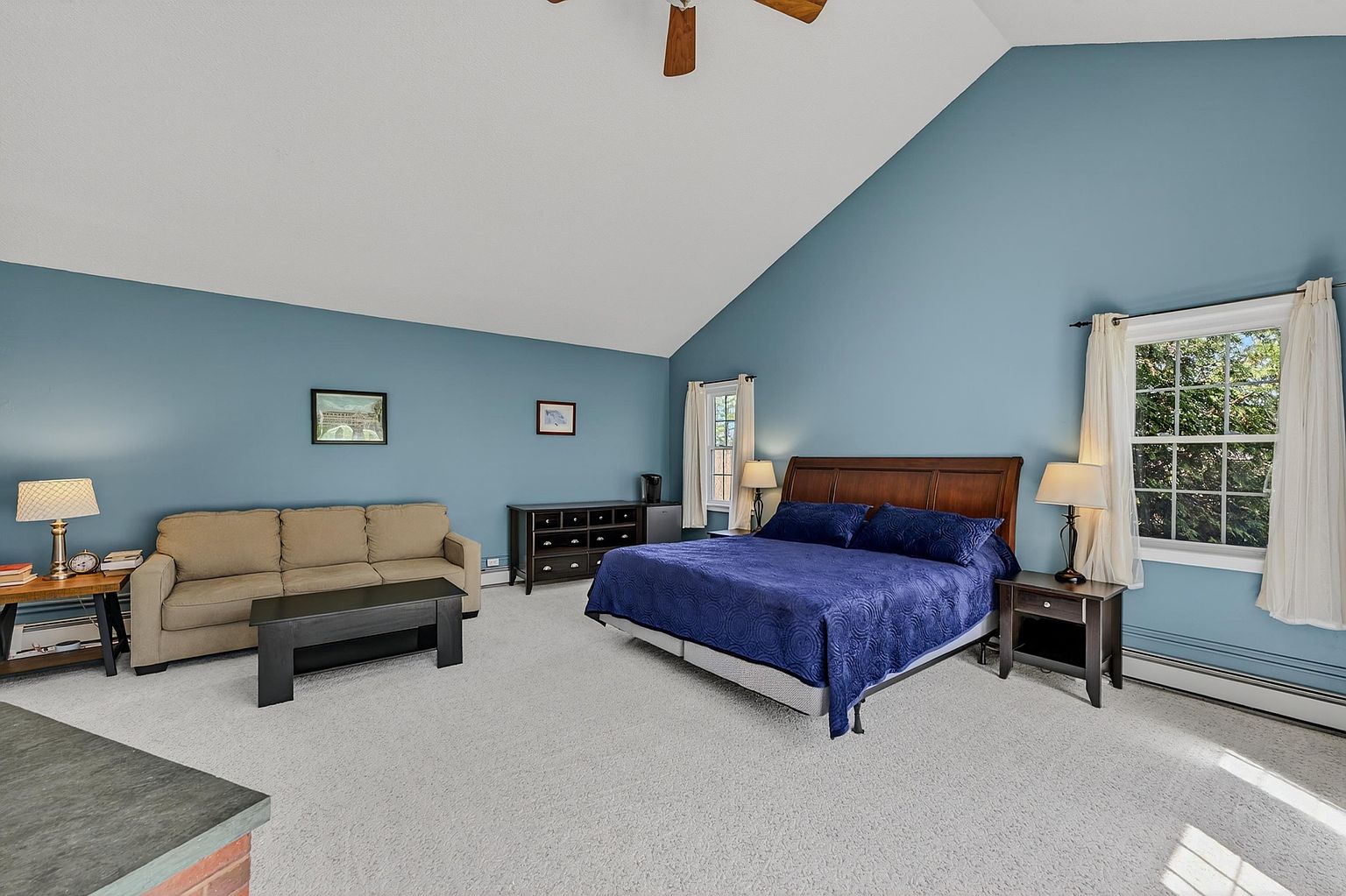 This spacious primary bedroom features a vaulted ceiling and soft blue walls, creating a calm and airy atmosphere. The room is furnished with a large wooden bed frame, a beige sofa, and a dark wood dresser, all set against a neutral carpet. Large windows with white curtains allow natural light to brighten the space, while bedside lamps provide warm accent lighting.