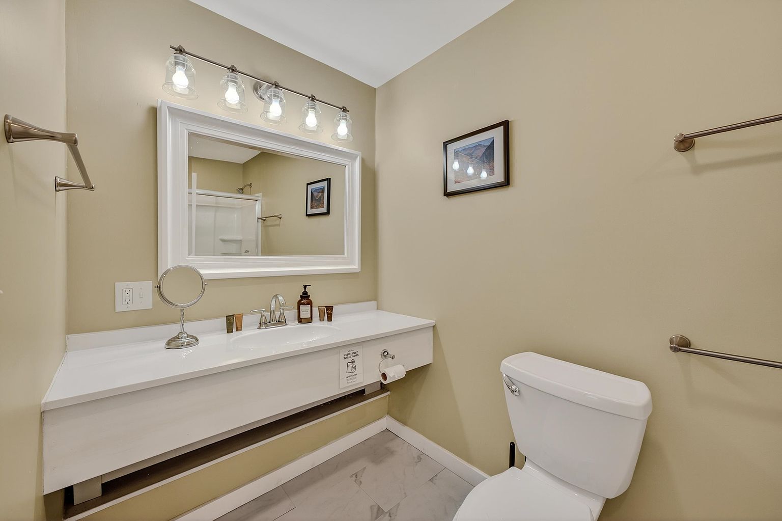 This clean and functional bathroom features a white vanity with a single sink, topped by a large, framed mirror and a multi-bulb light fixture. The walls are painted a soft neutral tone, complemented by light-colored tile flooring and a standard white toilet. A towel rack is mounted on the wall, and the space is accented by a small framed piece of art, creating a simple and well-maintained atmosphere.
