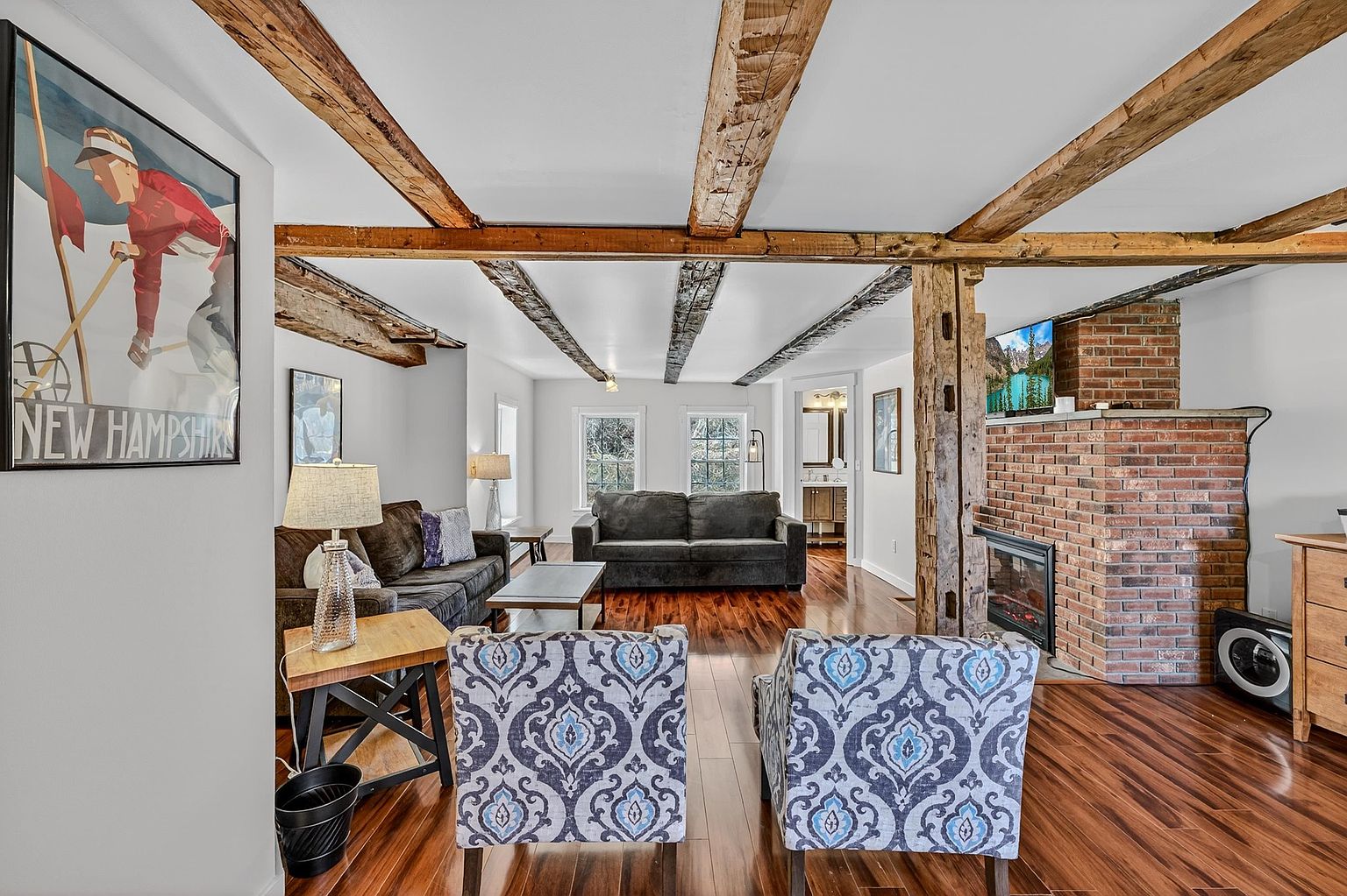 This inviting living room features rustic exposed wooden ceiling beams and rich, dark-toned hardwood flooring that runs throughout the space. The room is anchored by a brick chimney breast with an integrated fireplace and a television mounted above, while two patterned armchairs and a dark sofa provide comfortable seating. A large vintage-style New Hampshire poster on the wall adds a touch of local character to the warm, traditional aesthetic.