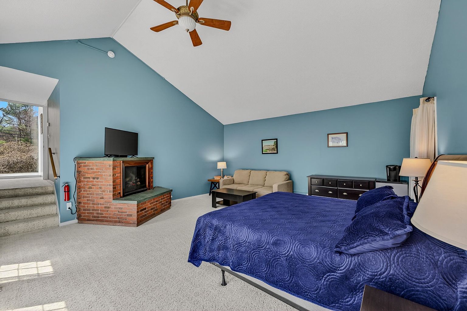 This spacious primary bedroom features a vaulted ceiling, light blue walls, and neutral carpeting throughout. The room is anchored by a large bed with a deep blue quilt and includes a brick fireplace with a television mounted above, a beige sofa, and a dark wood dresser. A set of stairs leads up to an exterior doorway, providing a unique architectural layout with ample natural light.