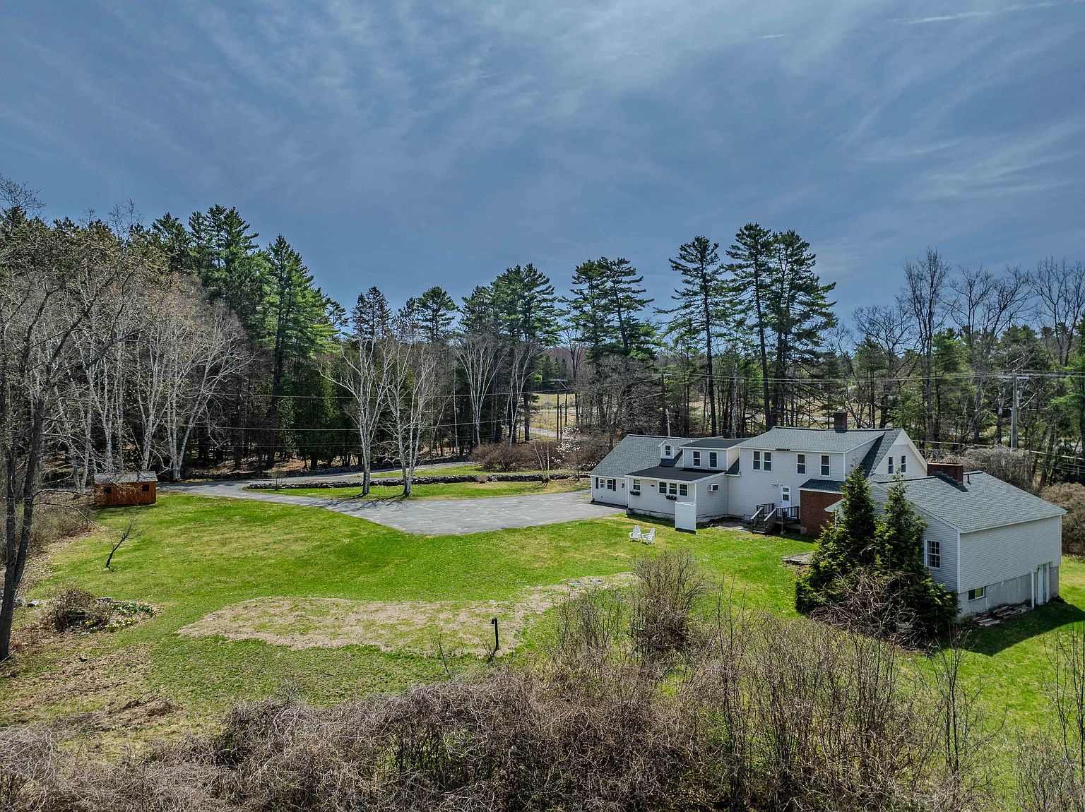 This aerial perspective captures a sprawling property featuring a multi-sectioned house with light-colored siding and a dark shingled roof. The home is set against a backdrop of dense, mature trees, with a large grassy lawn and a paved driveway area in the foreground. A small wooden shed sits to the left, and the overall setting offers a quiet, rural atmosphere with ample outdoor space.