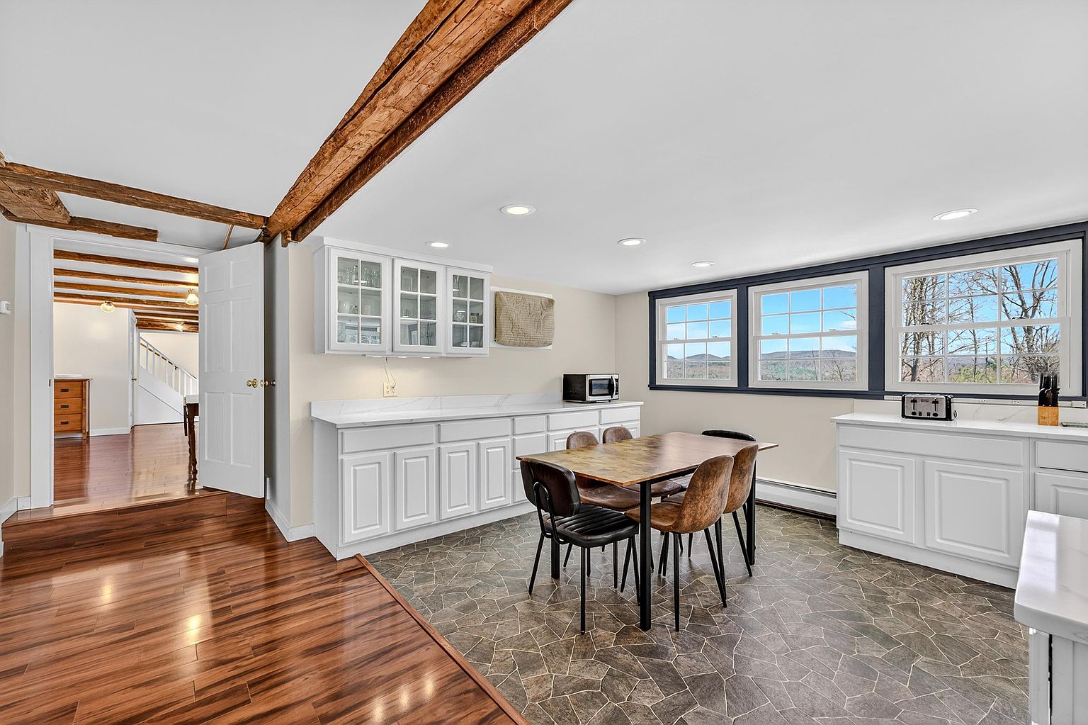 This bright kitchen features white cabinetry with glass-front upper units and light-colored countertops, providing a clean and functional workspace. A dining table with mixed-style chairs sits in the center, set against a backdrop of large windows that offer a scenic view of the outdoors. The room is defined by rustic exposed wooden ceiling beams and a transition between hardwood flooring and patterned stone-look tile.
