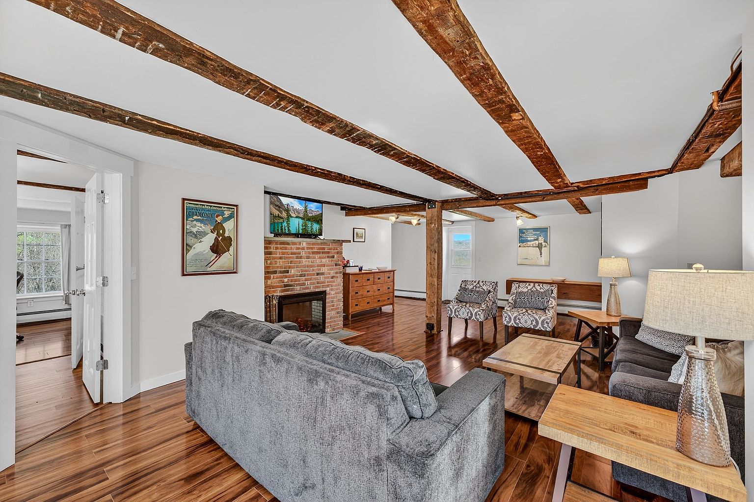 This spacious living room features exposed wooden ceiling beams and rich, warm-toned wood flooring, creating a rustic yet refined atmosphere. A brick chimney breast serves as a focal point, housing a fireplace and a television above, while two patterned armchairs and a plush gray sofa provide comfortable seating. The open-concept layout flows into an adjacent area, with a doorway leading to another room, all illuminated by soft, natural light.