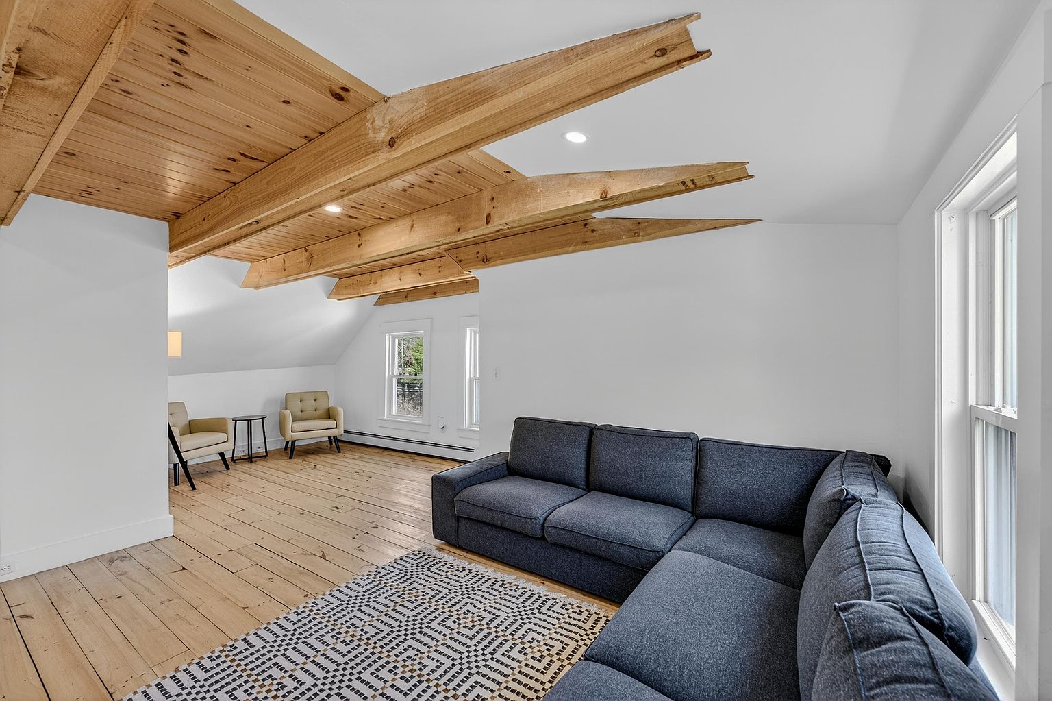 This spacious living area features a large, dark blue sectional sofa positioned on a patterned area rug, set against a backdrop of light wood flooring and white walls. The room is defined by its unique vaulted ceiling, which showcases exposed wooden beams and recessed lighting, creating a rustic yet modern aesthetic. A secondary seating area with two armchairs and a small side table is tucked into the corner, providing a cozy nook near the windows.