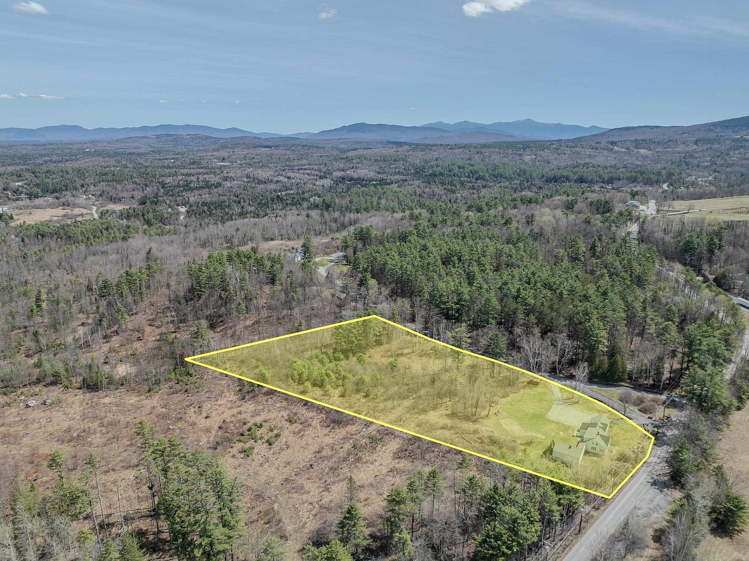 This high-angle aerial view captures a rural property highlighted by a yellow boundary overlay, showcasing a house situated on a large, partially wooded lot. The landscape features a mix of open grassy areas and dense forest, with a paved road running along the edge of the property. In the distance, rolling hills and mountain ranges provide a scenic backdrop under a clear blue sky.