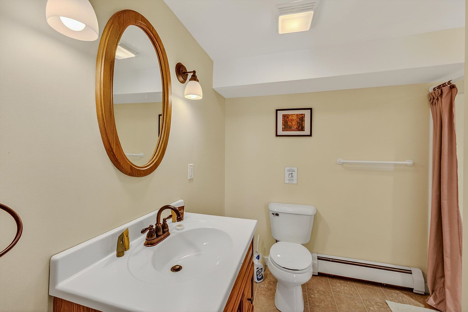 This bathroom features a vanity with a white sink and a warm-toned wood cabinet, complemented by a matching oval mirror and wall-mounted light fixture. The space includes a standard white toilet and a shower area partially obscured by a soft-toned curtain. The room is finished with light-colored walls and neutral flooring, creating a simple and functional atmosphere.