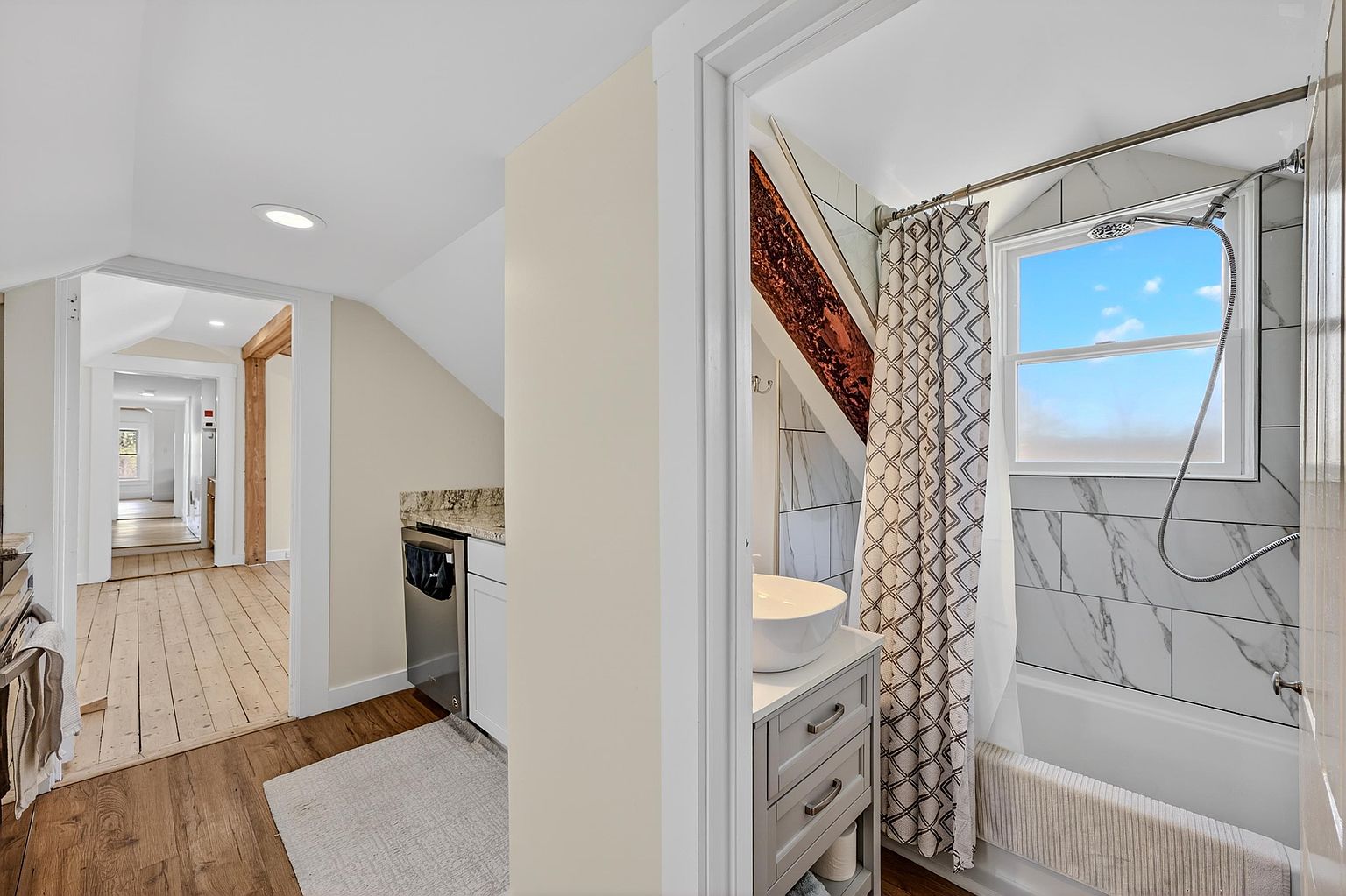 This view captures a compact bathroom featuring a vessel sink vanity and a tub-shower combination with light-colored patterned wall tiles. To the left, a doorway leads into a hallway with light wood flooring, revealing a glimpse of a kitchen area with a dishwasher and stone-topped counter. The space is characterized by its unique architectural angles, including a prominent exposed wooden beam, and a bright, airy atmosphere.