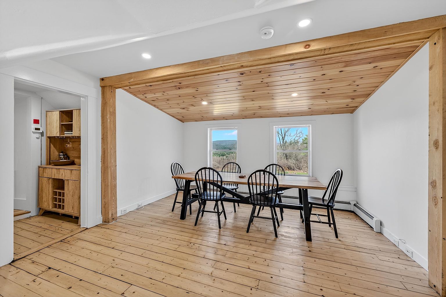 This dining room features a rustic aesthetic with light wood flooring and a matching wood-paneled ceiling. A long wooden table is surrounded by black spindle-back chairs, positioned beneath recessed lighting. The space is framed by exposed wooden beams and includes a view into an adjacent alcove with a wooden hutch.