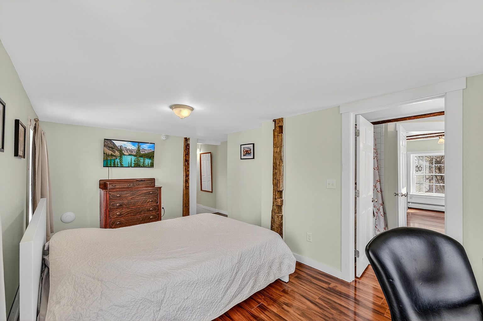 This bedroom features a neutral color palette with light-toned walls and warm-toned wood flooring. The room is anchored by a bed with a white headboard and a textured white coverlet, complemented by a dark wood dresser topped with a television. Two prominent, rustic exposed wooden support beams add architectural character, while a doorway leads to an adjacent room with a view of a window.