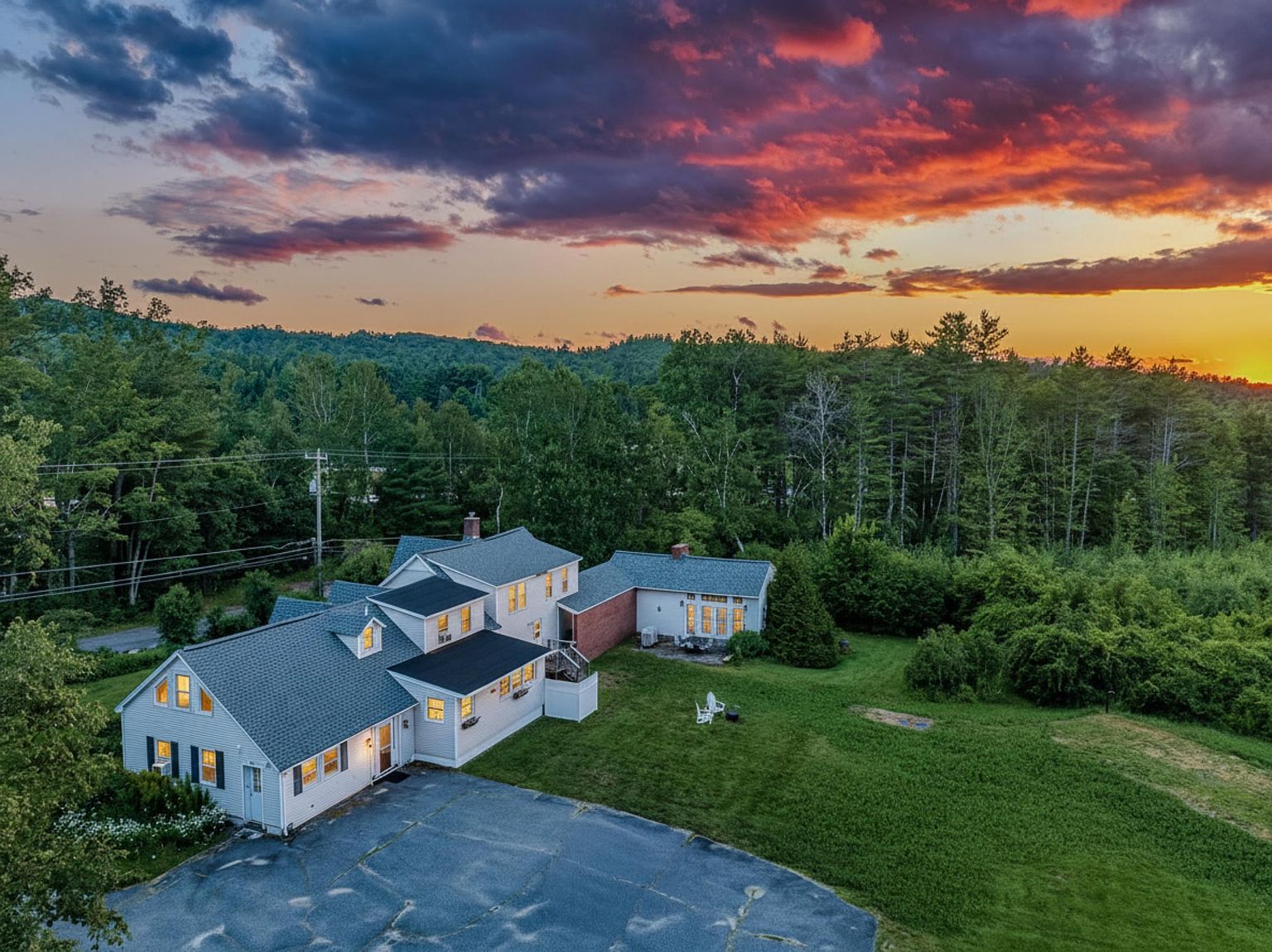 This aerial perspective captures a multi-sectioned residential property set against a vibrant, dramatic sunset sky. The home features light-colored siding, dark shingled roofs, and multiple dormer windows, with a paved driveway leading to the main entrance. A spacious green lawn extends from the house toward a dense backdrop of mature trees, creating a serene and picturesque setting.
