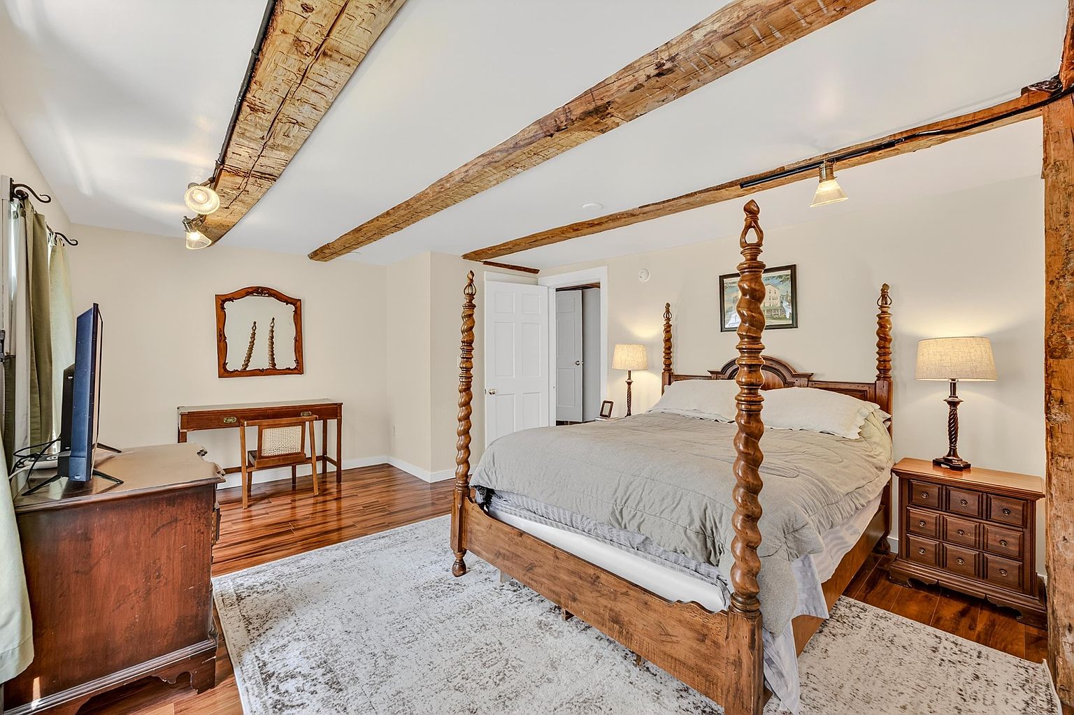 This spacious primary bedroom features a prominent four-poster wooden bed as the centerpiece, complemented by exposed wooden ceiling beams that add a rustic, historic charm. The room includes a dedicated desk area with a chair, a wooden dresser, and a nightstand, all set against neutral walls and warm-toned wood flooring. A large, light-colored area rug anchors the space, while track lighting provides adjustable illumination throughout the room.