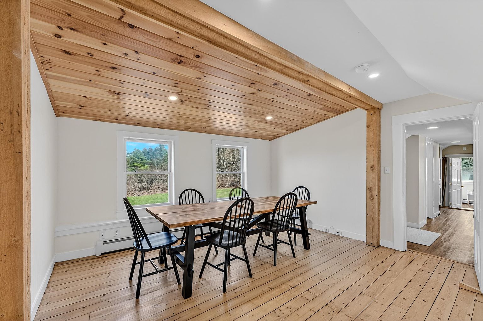 This dining room features a rustic aesthetic with light wood plank flooring and a matching wood-paneled ceiling. A large rectangular dining table with a natural wood top and black metal base is surrounded by six black spindle-back chairs. The space is brightened by two windows overlooking the outdoors and is framed by a prominent exposed timber post, creating an open transition into the adjacent hallway.