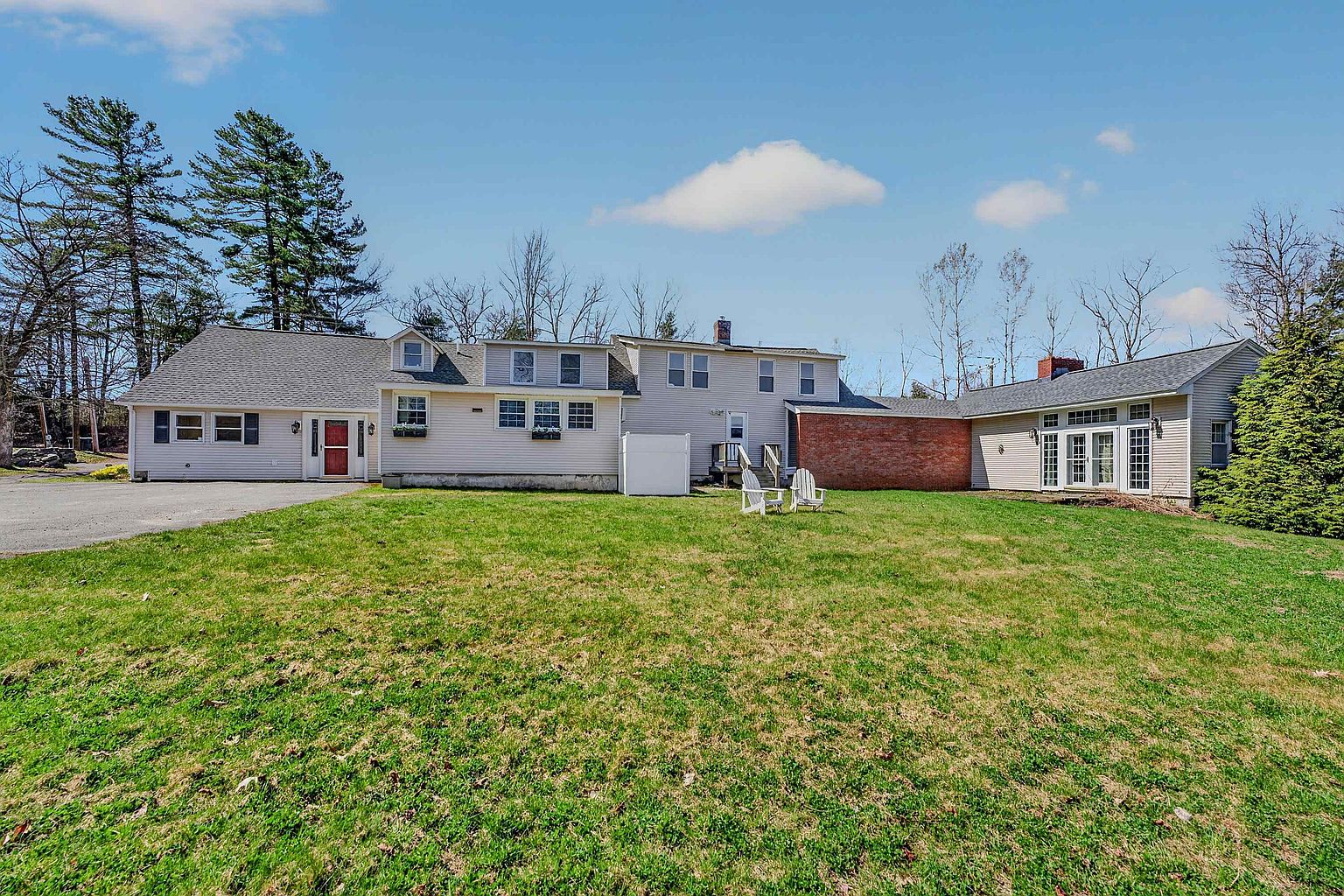This rear view of a multi-sectioned residential property showcases a sprawling lawn leading up to a light-colored siding exterior. The home features a mix of architectural rooflines, a prominent brick section, and multiple windows, with a pair of white Adirondack chairs placed on the grass. The perspective is a wide, eye-level shot that captures the expansive backyard and the full length of the house under a bright, clear blue sky.