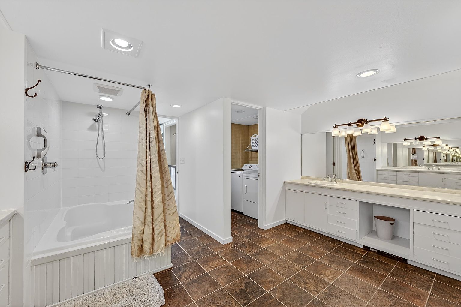 This spacious primary bathroom features a combination tub and shower unit with white tile surround and a beige curtain. To the right, a long vanity with a single sink and a large wall-to-wall mirror provides ample counter space, while a doorway leads to a separate laundry area. The room is finished with dark, earth-toned floor tiles and recessed lighting, creating a functional and neutral aesthetic.