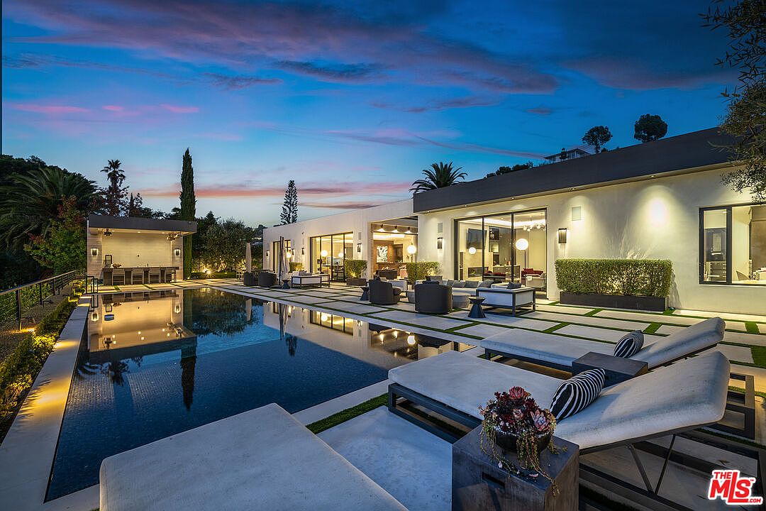 This image showcases a luxurious backyard with a stunning pool and spa area. The modern home features large glass doors that open to an outdoor living space with comfortable seating. The pool reflects the ambient lighting and the sky, creating a serene and inviting atmosphere, while lounge chairs are placed near the pool for relaxation.