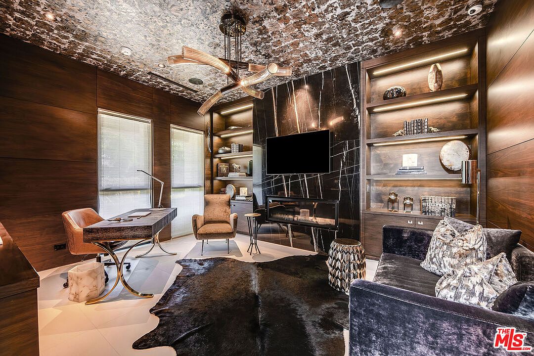This is a luxurious home office featuring dark wood paneling, a textured ceiling, and a black marble accent wall with a mounted television. The room includes built-in shelving with accent lighting, a modern desk with a leather chair, and a comfortable seating area with a dark velvet sofa and decorative pillows. A cowhide rug adds texture to the space, and a unique chandelier hangs from the ceiling, creating a sophisticated and professional atmosphere.