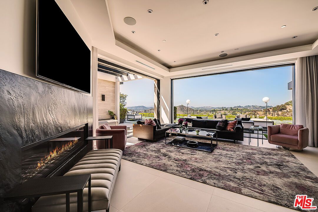 This is a luxurious living room featuring a modern fireplace with a large television above it. The room has an open floor plan with expansive windows offering stunning views of the landscape. The decor includes stylish furniture, a patterned rug, and elegant lighting, creating a sophisticated and inviting atmosphere.
