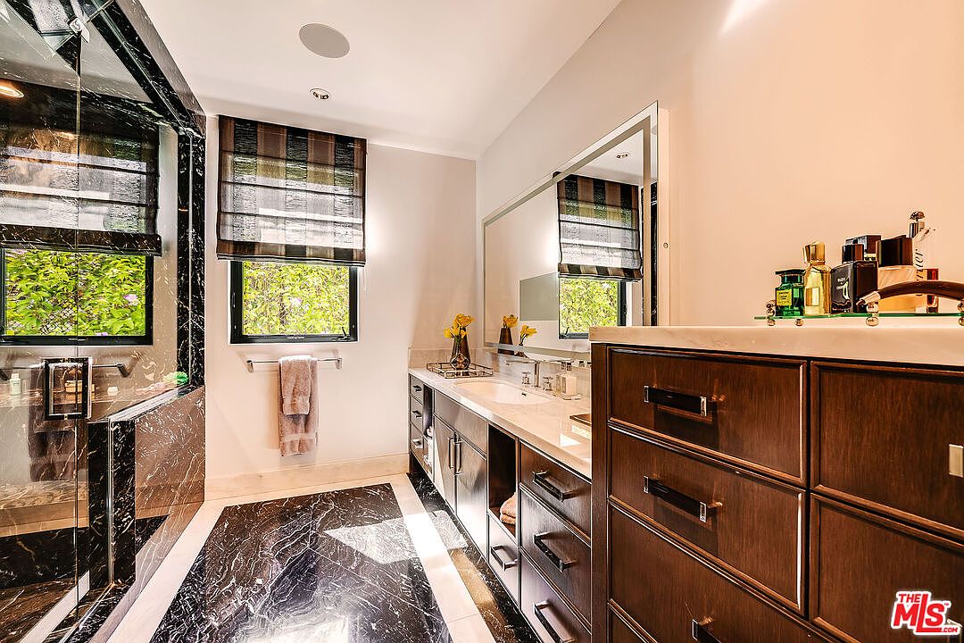 This is a primary bathroom featuring a black and white marble floor and shower. The vanity has dark wood cabinets and a white countertop with dual sinks. A large mirror hangs above the vanity, and there are windows with striped shades.