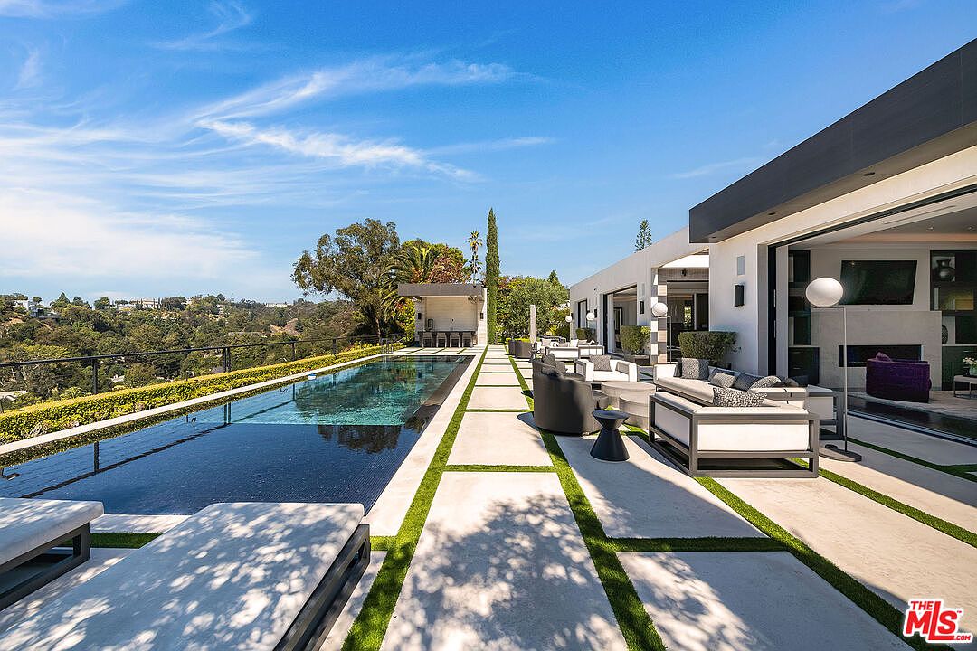 This image showcases a luxurious outdoor space featuring a long, sleek swimming pool with an infinity edge, surrounded by modern patio furniture and lush landscaping. The pool area seamlessly transitions into an open-air living space with comfortable seating, creating an inviting atmosphere for relaxation and entertainment. The architectural style is contemporary, with clean lines and a focus on indoor-outdoor living.