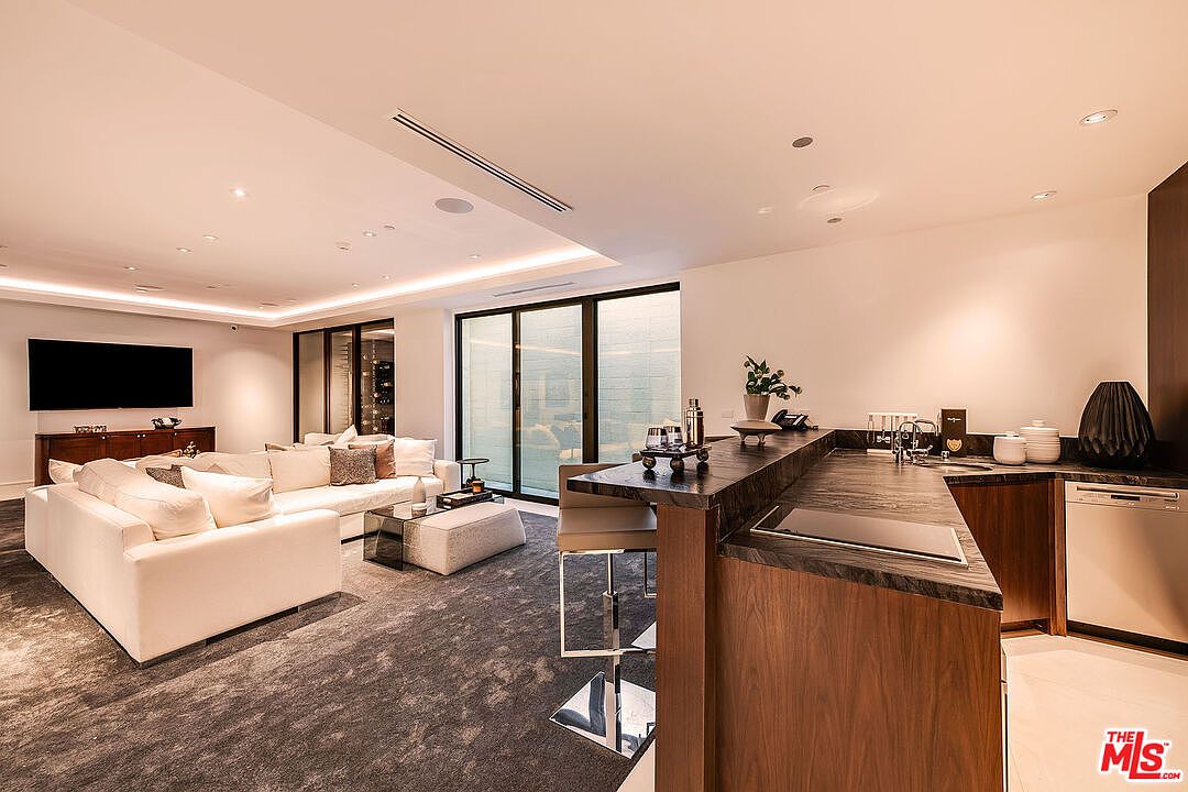This interior shot showcases a modern living room and kitchen area. The living room features a large white sofa, a glass coffee table, and a dark gray rug, while the kitchen area includes a dark countertop bar with seating and modern appliances. The room is well-lit with recessed lighting and natural light from the large windows, creating a luxurious and inviting atmosphere.