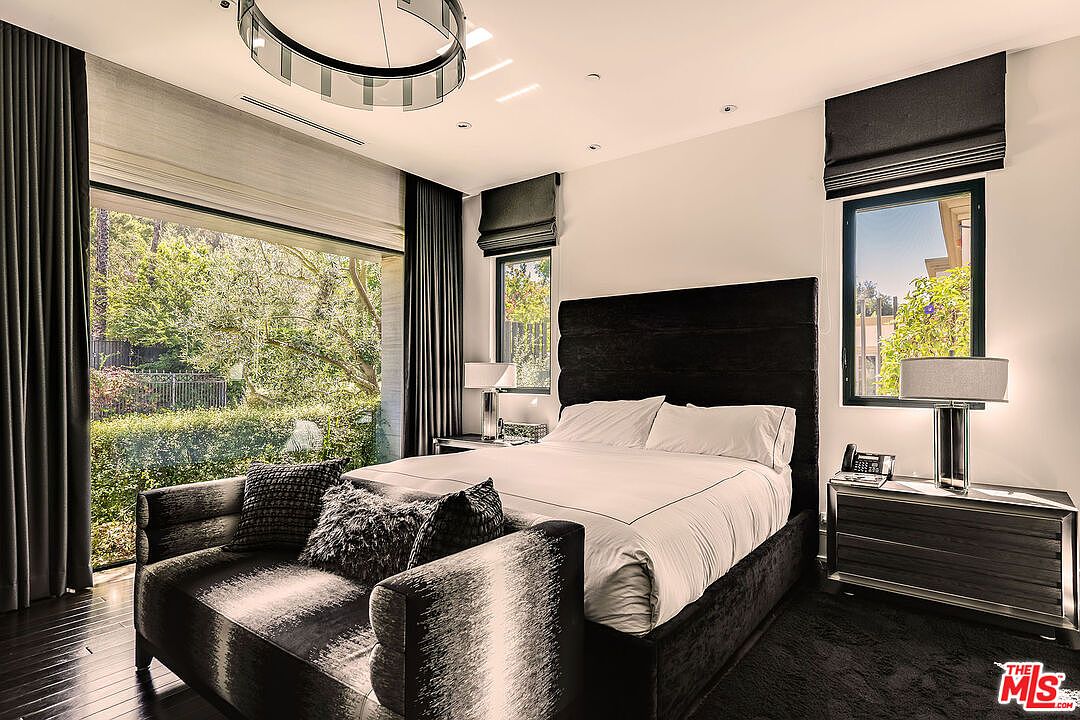 This is a luxurious primary bedroom featuring a large bed with a dark upholstered headboard and crisp white linens. A stylish chaise lounge sits at the foot of the bed, adorned with decorative pillows. Large windows offer views of lush greenery, and modern lighting fixtures add to the room's sophisticated ambiance.