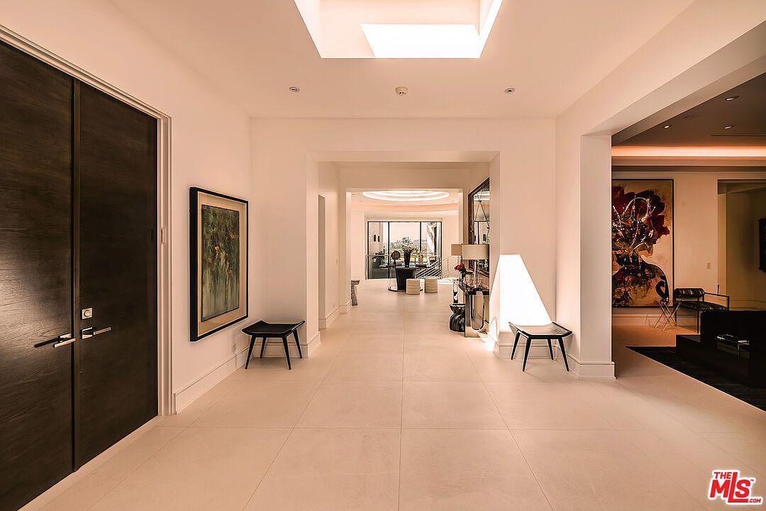 This is an interior shot of a modern hallway featuring light tile flooring and white walls. The hallway is well-lit with a skylight and recessed lighting, leading to other rooms visible through archways. The space is decorated with artwork and minimalist furniture, creating a clean and sophisticated aesthetic.