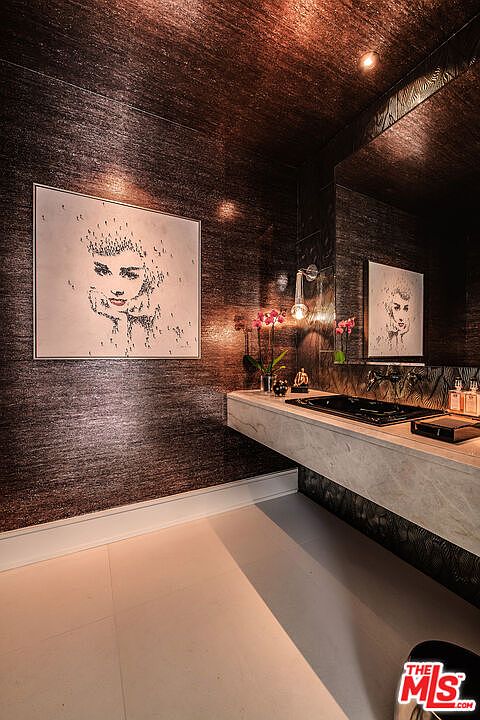 This is a stylish guest bathroom featuring dark, textured wallpaper and a floating vanity with a light-colored countertop. A large framed portrait hangs on the wall, complemented by a smaller one reflected in the mirror. The room is illuminated by a modern wall sconce, creating a sophisticated and luxurious ambiance.