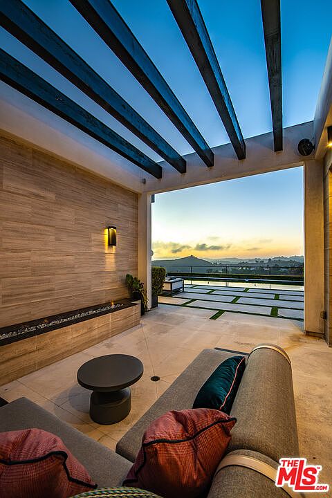 This image showcases a luxurious outdoor patio area with a modern design. The space features comfortable lounge seating with decorative pillows, a sleek side table, and a built-in water feature along a textured wall. An open pergola structure provides partial shade, while the patio overlooks a stunning view of the landscape and a pool area, creating an inviting and relaxing atmosphere.