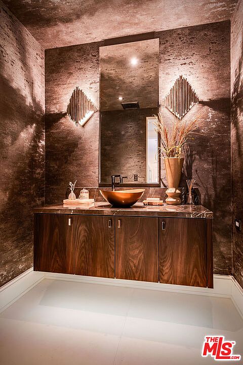This is a modern bathroom featuring a floating wood vanity with a copper vessel sink and a sleek black faucet. A large mirror hangs above the vanity, flanked by unique diamond-shaped sconces. The walls are covered in a textured, dark-toned material, and the floor is a light tile, creating a sophisticated and luxurious ambiance.