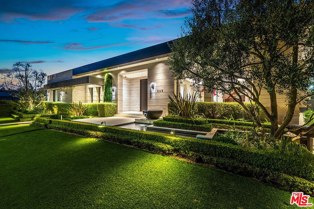 This is a front exterior view of a modern home at dusk, showcasing its sleek architectural design and meticulously landscaped front yard. The property features a water feature, manicured hedges, and ambient lighting that highlights the facade and lush greenery. The overall impression is one of sophisticated elegance and curb appeal.