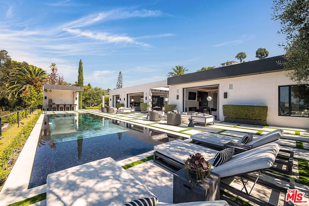 This image showcases a luxurious backyard with a stunning pool and modern architectural design. The pool features dark tiles and is surrounded by sleek lounge chairs and manicured landscaping. The contemporary home boasts large sliding glass doors that open to the outdoor living space, creating a seamless indoor-outdoor flow.