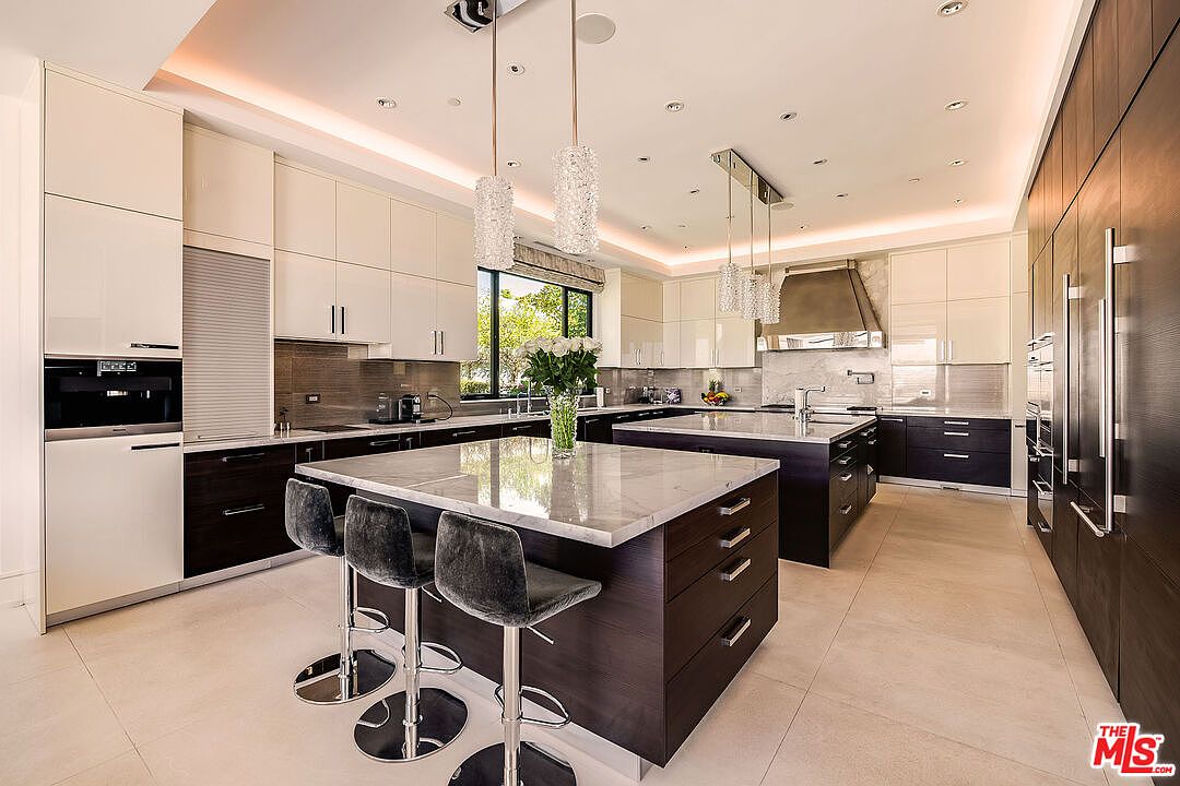 This is a modern kitchen with two islands, white and dark wood cabinetry, and stainless steel appliances. The kitchen features pendant lighting, marble countertops, and a window overlooking the outdoors. The overall impression is luxurious and spacious, perfect for a high-end real estate listing.