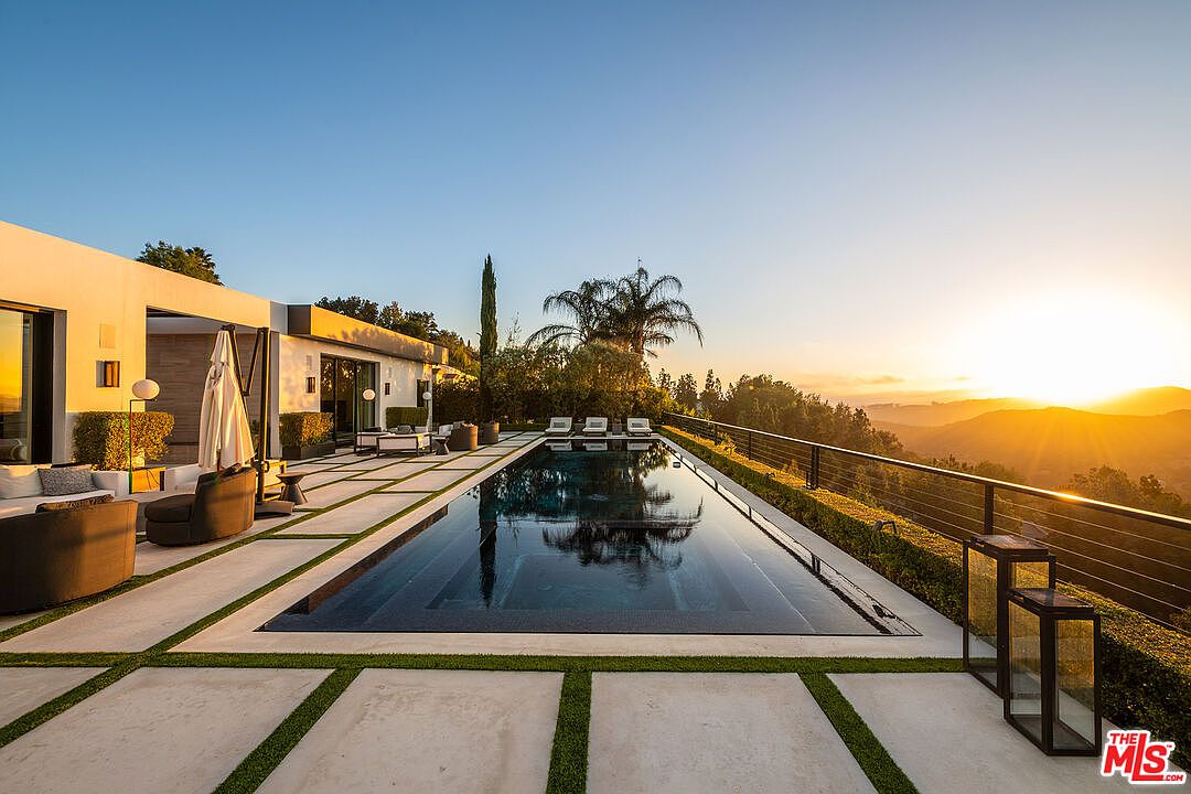 This image showcases a luxurious infinity pool at sunset, reflecting the sky and surrounding landscape. The pool is surrounded by a modern patio with lounge seating and manicured greenery, offering a serene and private outdoor space. A sleek railing provides safety without obstructing the stunning panoramic views of the hills and setting sun.