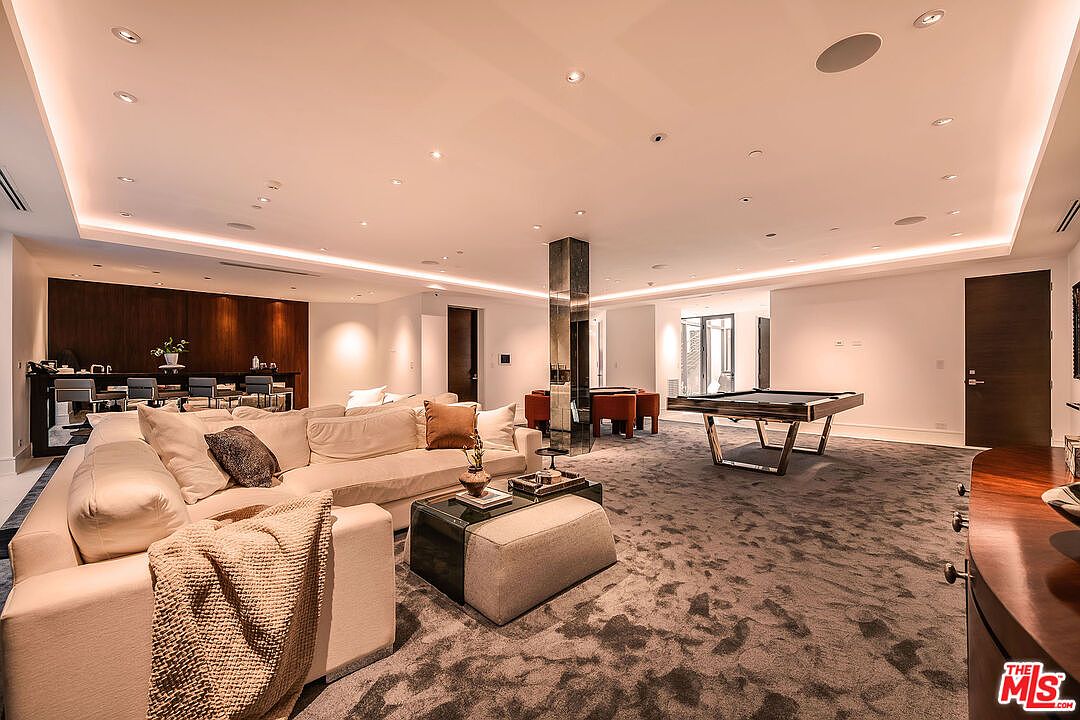 This is a spacious living room featuring a large sectional sofa with throw pillows and a blanket, a glass-topped coffee table with an ottoman, and a pool table. The room has a neutral color palette with gray carpeting and white walls, accented by a dark wood feature wall and a mirrored column. Recessed lighting provides ample illumination, creating a bright and inviting atmosphere.