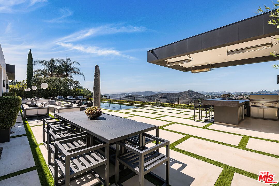 This image showcases a luxurious outdoor patio area with a modern design. The space features a large dining table with chairs, a built-in outdoor kitchen with a grill, and a lounge area with comfortable seating. The patio is surrounded by a well-maintained landscape and offers stunning views of the surrounding mountains and cityscape, creating an inviting and upscale atmosphere.