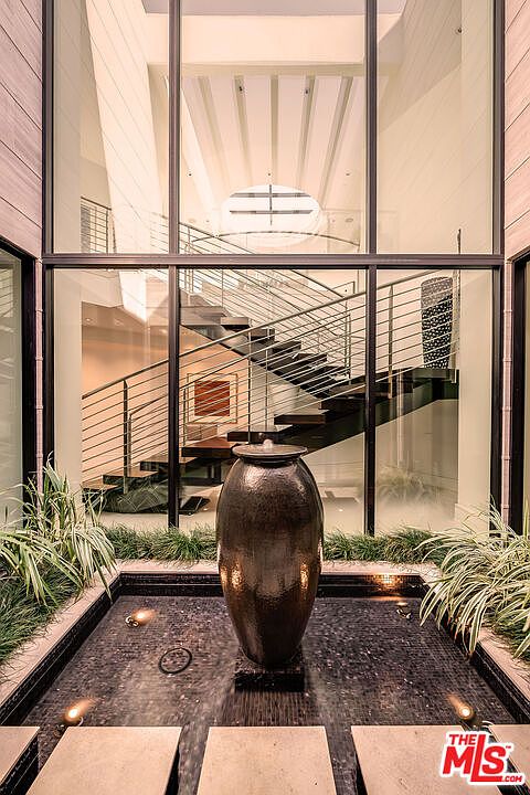 This image showcases a striking architectural feature: a modern indoor water feature set against a backdrop of a contemporary staircase visible through large windows. The water feature includes a dark, urn-shaped fountain in a shallow pool, surrounded by greenery and stone pavers. The overall impression is one of tranquility and sophisticated design, highlighting the home's unique and luxurious elements.