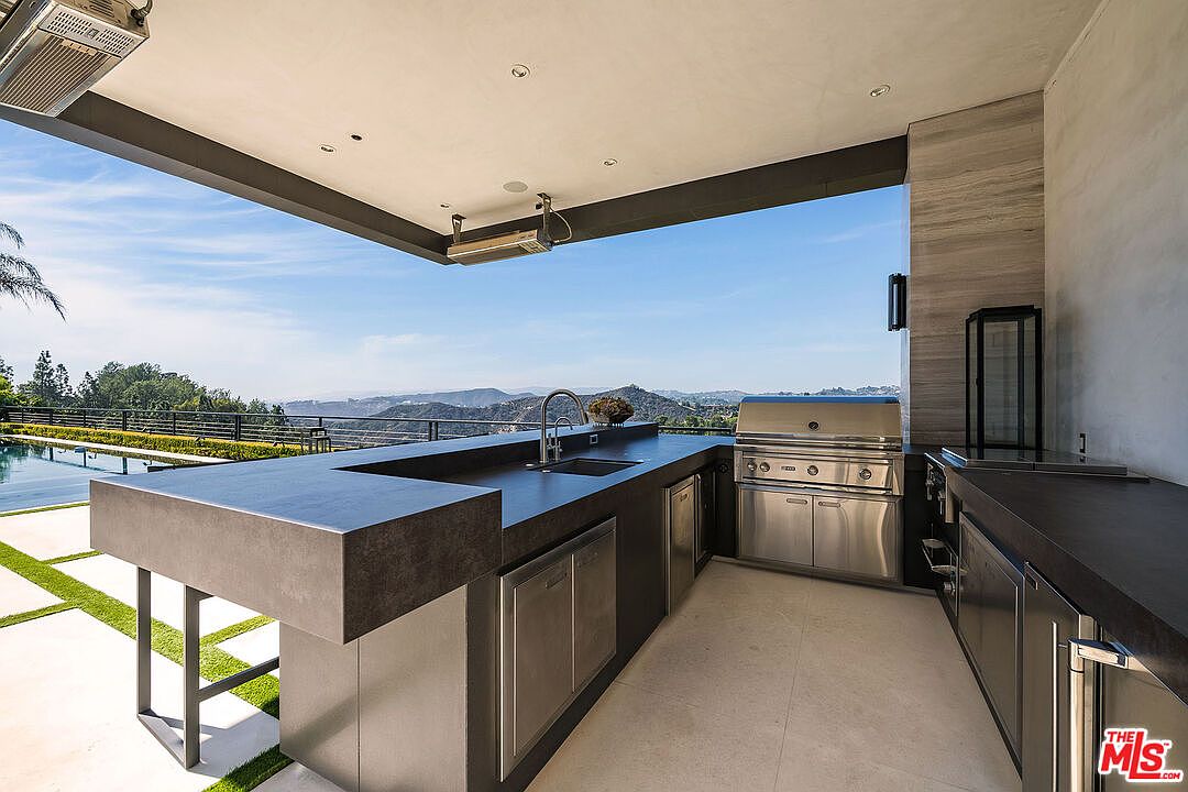 This image showcases a luxurious outdoor kitchen and patio area with stunning panoramic views. The kitchen features a built-in grill, sleek countertops, and stainless steel appliances, perfect for outdoor entertaining. The patio is designed with modern aesthetics, offering a seamless blend of indoor and outdoor living with a view of the mountains and pool.