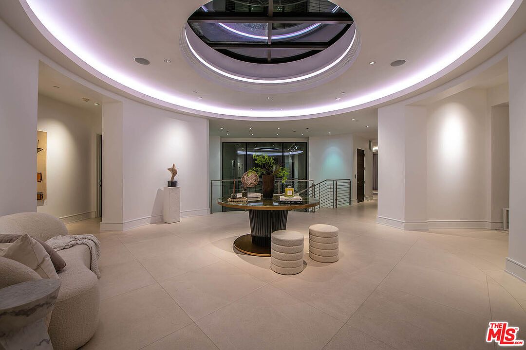 This interior shot showcases a luxurious hallway with a striking circular skylight, illuminating the space with natural light. A round table with decorative items and two cushioned stools anchor the center, while the neutral color palette and clean lines contribute to a modern and elegant aesthetic. A glimpse of stairs with metal railings can be seen in the background.